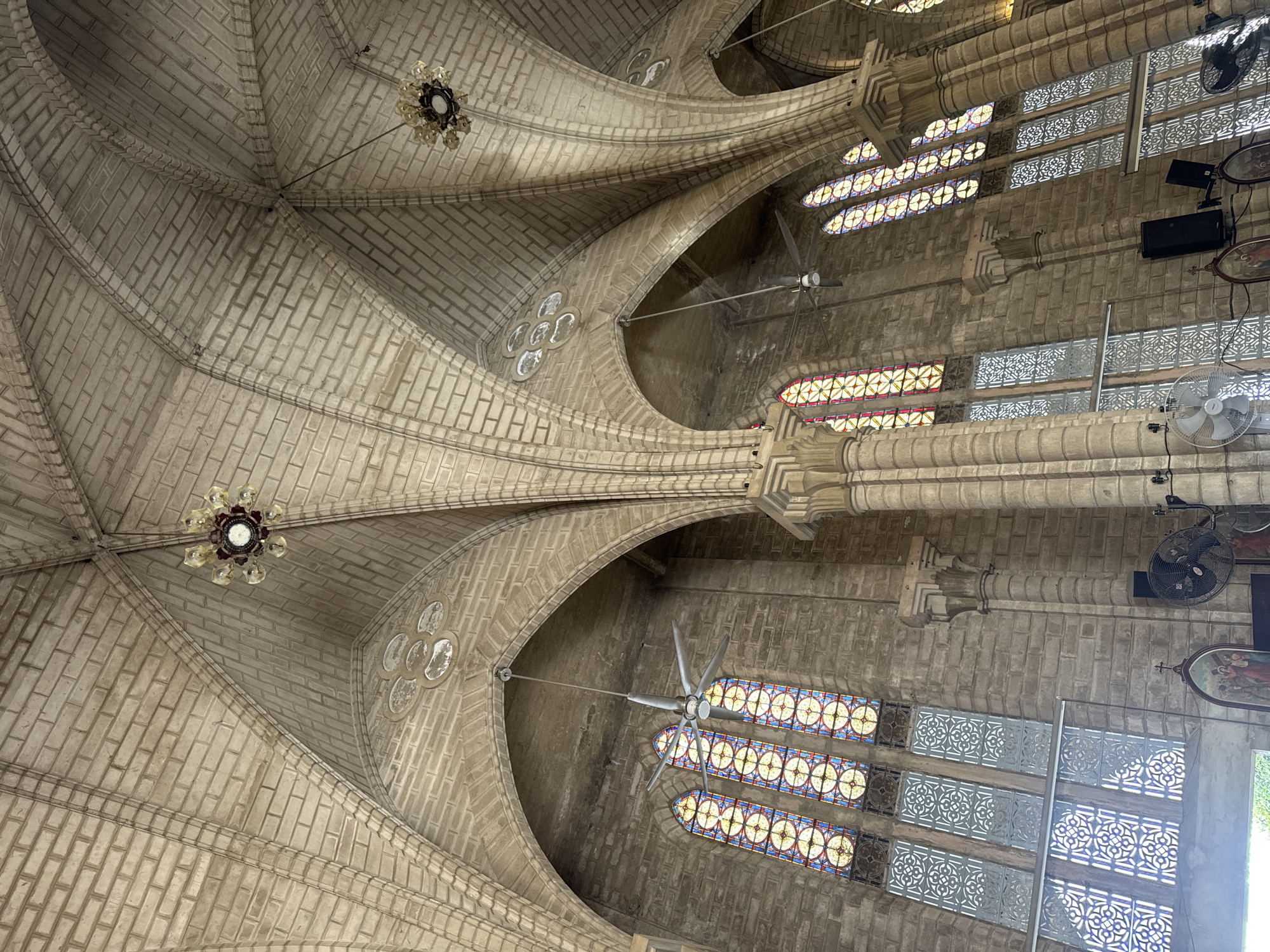The image shows the interior of Nha Trang Cathedral, a Roman Catholic cathedral in Nha Trang, Vietnam. The photograph is taken from a low angle, looking up at the vaulted ceiling and stained-glass windows. The architecture is Gothic-inspired, with pointed arches and ribbed vaults made of rough-hewn stone blocks. Several large ceiling fans are visible, along with ornate chandeliers. The stained-glass windows are tall and narrow, featuring geometric patterns and colorful designs. On the lower walls, decorative grilles and framed religious artwork are present. The overall impression is one of grandeur and historical significance, with a stark contrast between the ancient stone structure and the modern ceiling fans.