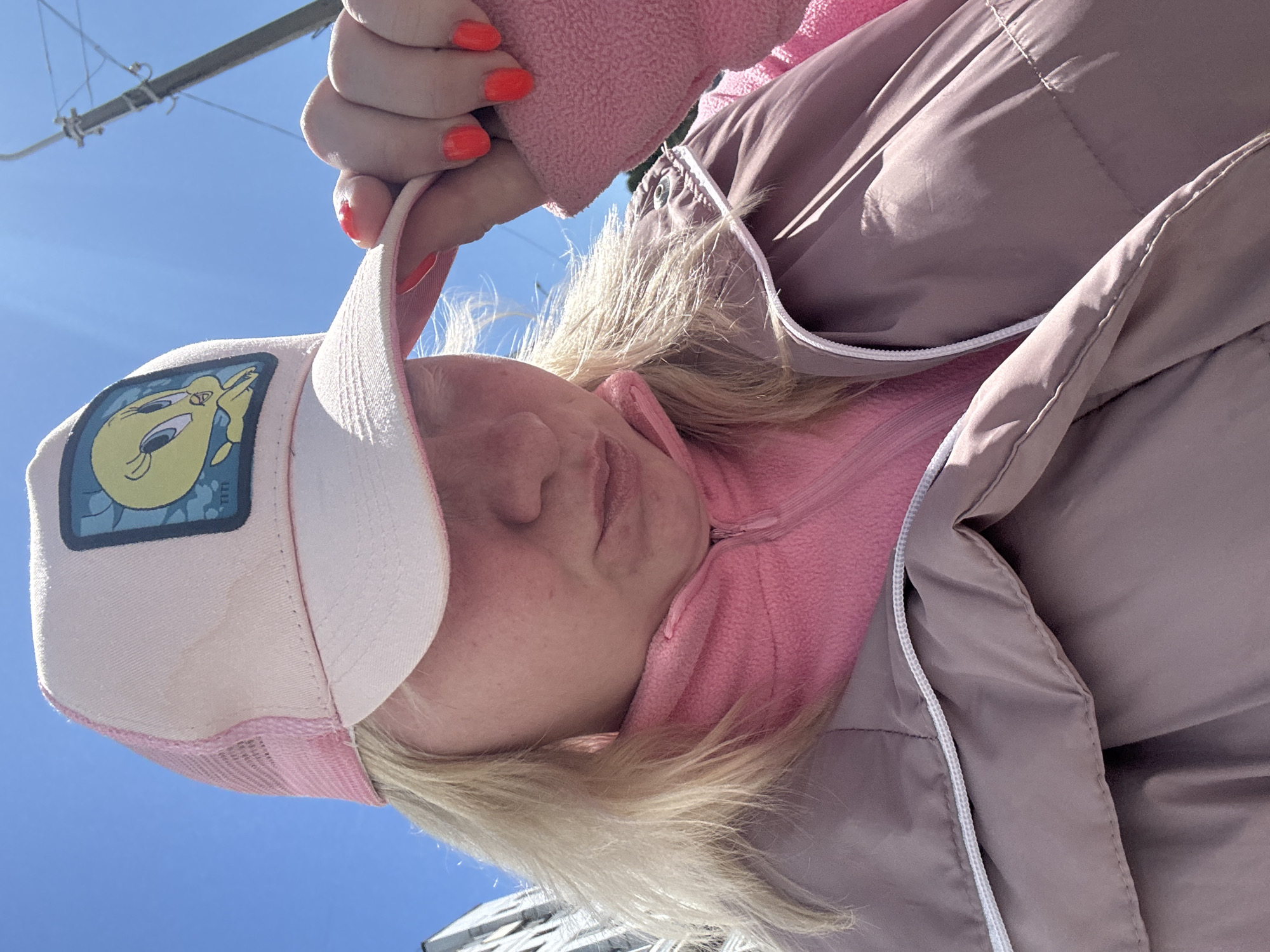 A woman is wearing a pink jacket, pink fleece, and pink trucker hat with Tweety Bird. The photo is an upward angle of the woman's face. The woman has blonde hair and red fingernails. In the background is a blue sky and the top of a building. A pole and wires are visible in the upper left corner. The location is Chaikovsky, Russia.