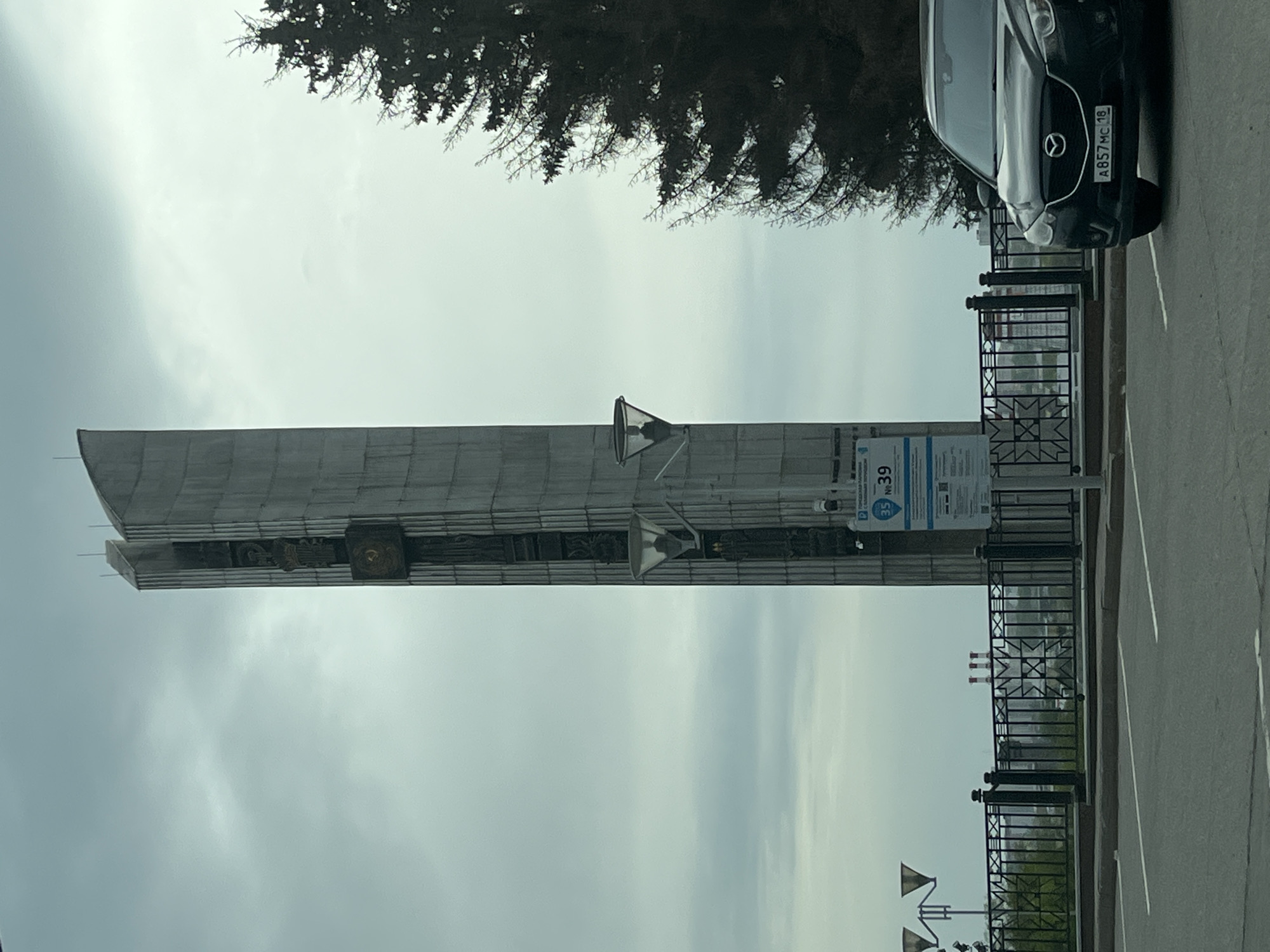 The image shows the "Forever with Russia" Monument in Izhevsk, Russia. The monument is a tall, vertical concrete structure adorned with sculpted elements. A black car is parked adjacent to a fence near the monument. The sky is overcast, and a portion of a tree is visible in the upper part of the frame.
