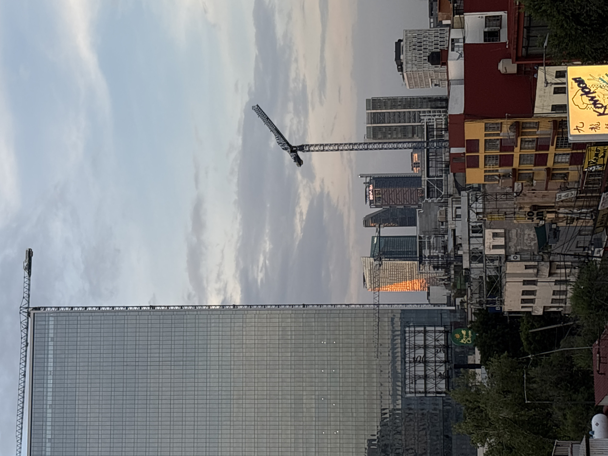 The image depicts a daytime view of a cityscape in Mexico City, Mexico. The sky is overcast with light-colored clouds. A tall, mirrored skyscraper dominates the foreground. Atop this building is a smaller crane-like structure. In the midground, several other skyscrapers are visible, along with a large construction crane. Closer to the right side of the image, a mix of older buildings and urban structures is visible, including a building with red and yellow accents and a neon sign with foreign text. Some trees are visible in the lower right corner. The overall impression is of a dense urban environment with ongoing construction and a mix of architectural styles.