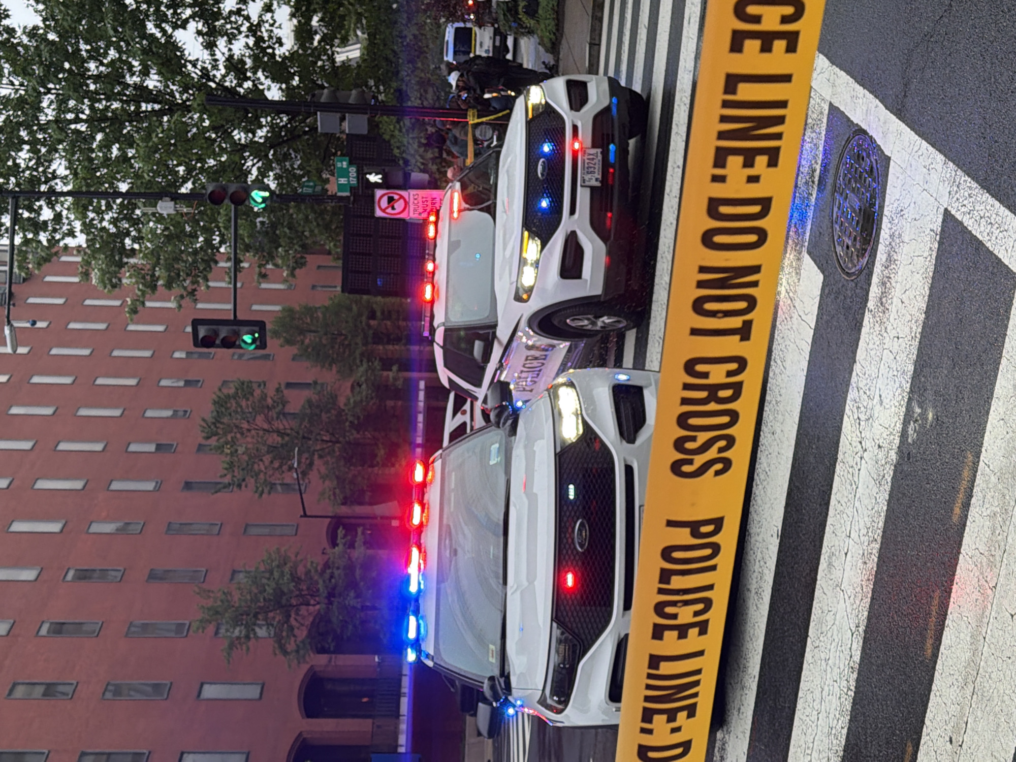 The image shows a street scene at night in the 1700 block of H Street in Washington, D.C., United States of America. Two white police vehicles with flashing red and blue lights are parked on the street, partially blocking a crosswalk. Yellow police tape reading "POLICE LINE: DO NOT CROSS POLICE LINE" is strung across the crosswalk. A "NO TRUCKS" sign is visible. A manhole cover is also visible in the crosswalk. A red brick building lines the left side of the street. The traffic lights are green. Several people are standing around the police vehicles.
