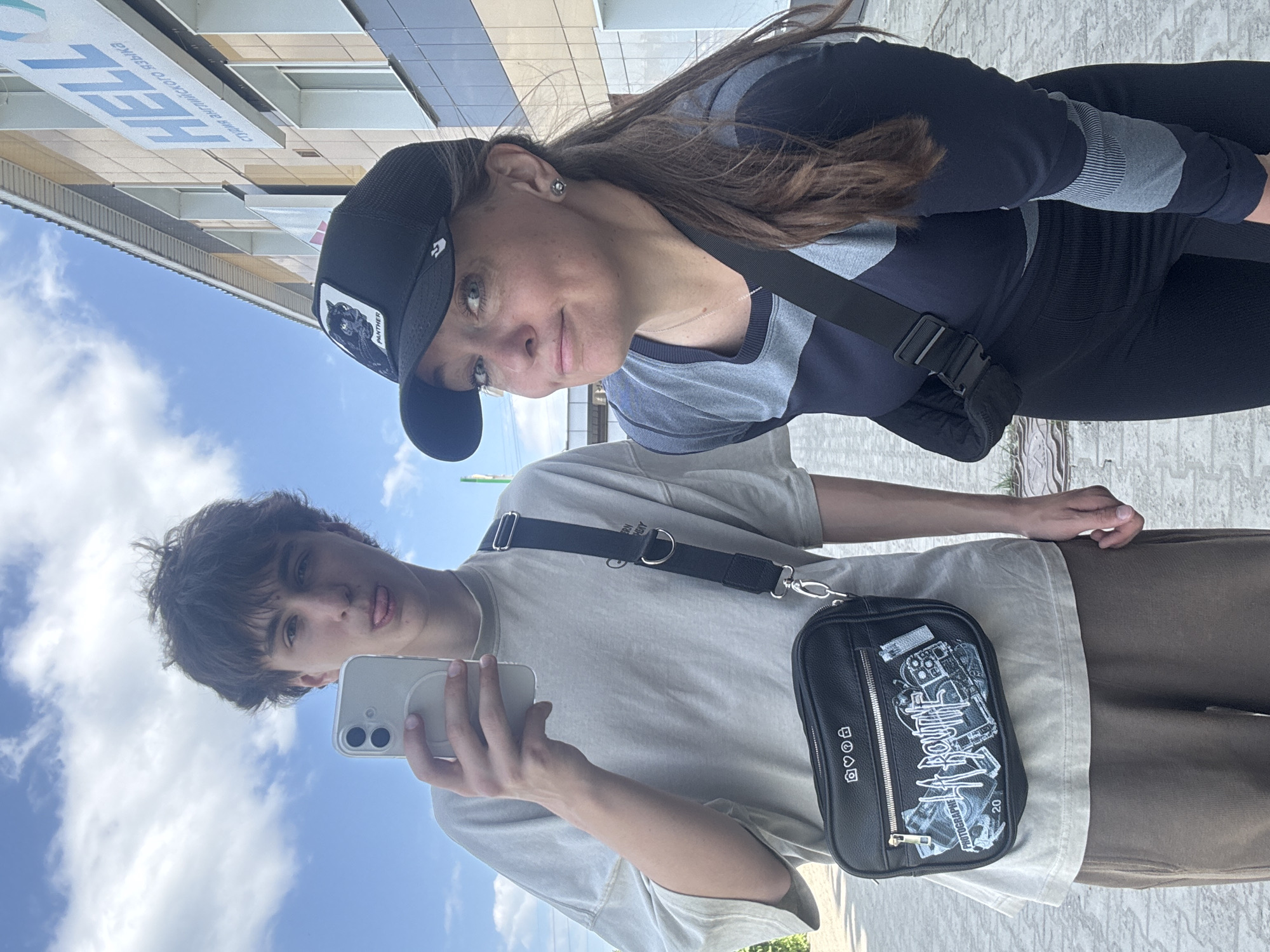 A young man and woman stand outside a building in the city of Chaikovsky, Russia. The woman wears a black baseball cap with a panther on it, a grey and blue top, and black leggings. She has a black purse strap across her chest. The man wears a white t-shirt, brown pants, and a black purse across his chest. He holds a white smartphone. The building has the word "HEL" written on it. The sky is blue with some clouds.
