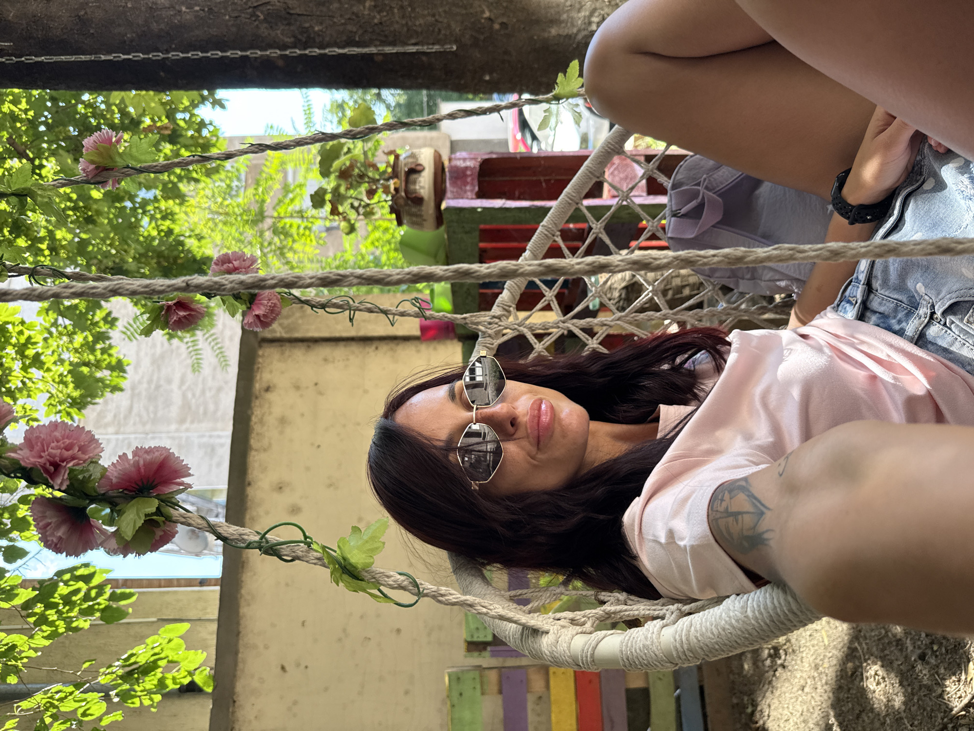 A woman is sitting in a white macrame hanging chair, outdoors. She has dark hair, is wearing sunglasses, a light pink t-shirt, and denim shorts. There is a tattoo on her left arm and a black watch on her left wrist. A light gray backpack is visible behind her. The chair is decorated with artificial pink flowers and green leaves. In the background, there is a tree with green foliage, a wall, and a colorful pallet bench. The scene suggests a casual, outdoor setting, possibly in a courtyard or garden area. The woman appears relaxed, looking towards the camera. The location in the city of Pleven, Bulgaria is not specified in the image itself.