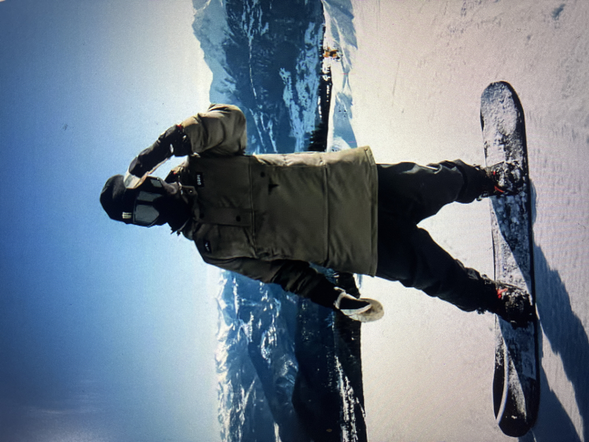 A male snowboarder stands on a snowy mountain slope under a clear blue sky. The image is oriented as if the snowboarder is upright, with the vast sky above and snow-covered mountains in the distance.
The snowboarder wears full winter attire, including a thick, olive green or khaki-colored winter jacket, dark snow pants, and black gloves. His face is largely obscured by a black balaclava or face mask, and he wears dark goggles. His left hand is raised towards his head, possibly adjusting his goggles or shielding his eyes from the sun. He is standing on a black snowboard, which is partially covered in snow.
The background features rugged, snow-capped mountains under a bright, cloudless blue sky, suggesting a high-altitude winter landscape. The ground around the snowboarder is covered in white snow. The scene depicts a clear, sunny winter day, likely morning or early afternoon.
On the left chest of the snowboarder's jacket, a small rectangular patch with light text is visible, which appears to read "GRPI" or a similar acronym. The overall impression is one of a solitary figure enjoying the serene and majestic mountain environment, perhaps pausing before or after a ride. The context provided indicates the location is near Asenovgrad, Bulgaria.