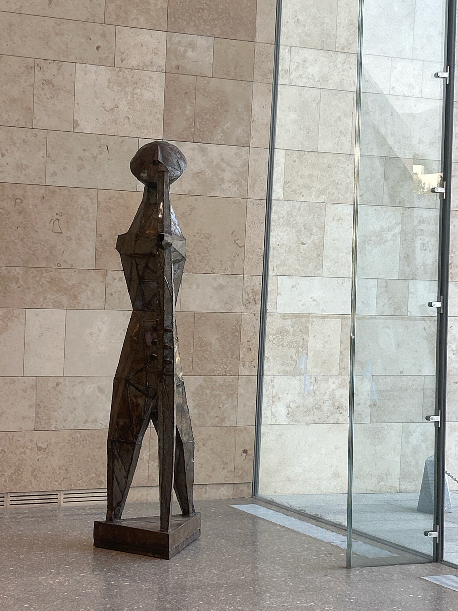 An abstract metal sculpture stands in a modern interior space. The sculpture is dark brown, elongated, and geometric, with a rounded head and thin, angular limbs. It is mounted on a dark rectangular base. The sculpture is positioned on a speckled gray floor. Behind the sculpture is a wall clad in beige, rectangular stone tiles. To the right of the sculpture is a large glass partition, through which a brightly lit outdoor area is partially visible, including what appears to be a stone structure. The scene suggests an indoor art exhibition or a public building with modern architectural features. The image's metadata indicates it was taken in Almaty, Kazakhstan.