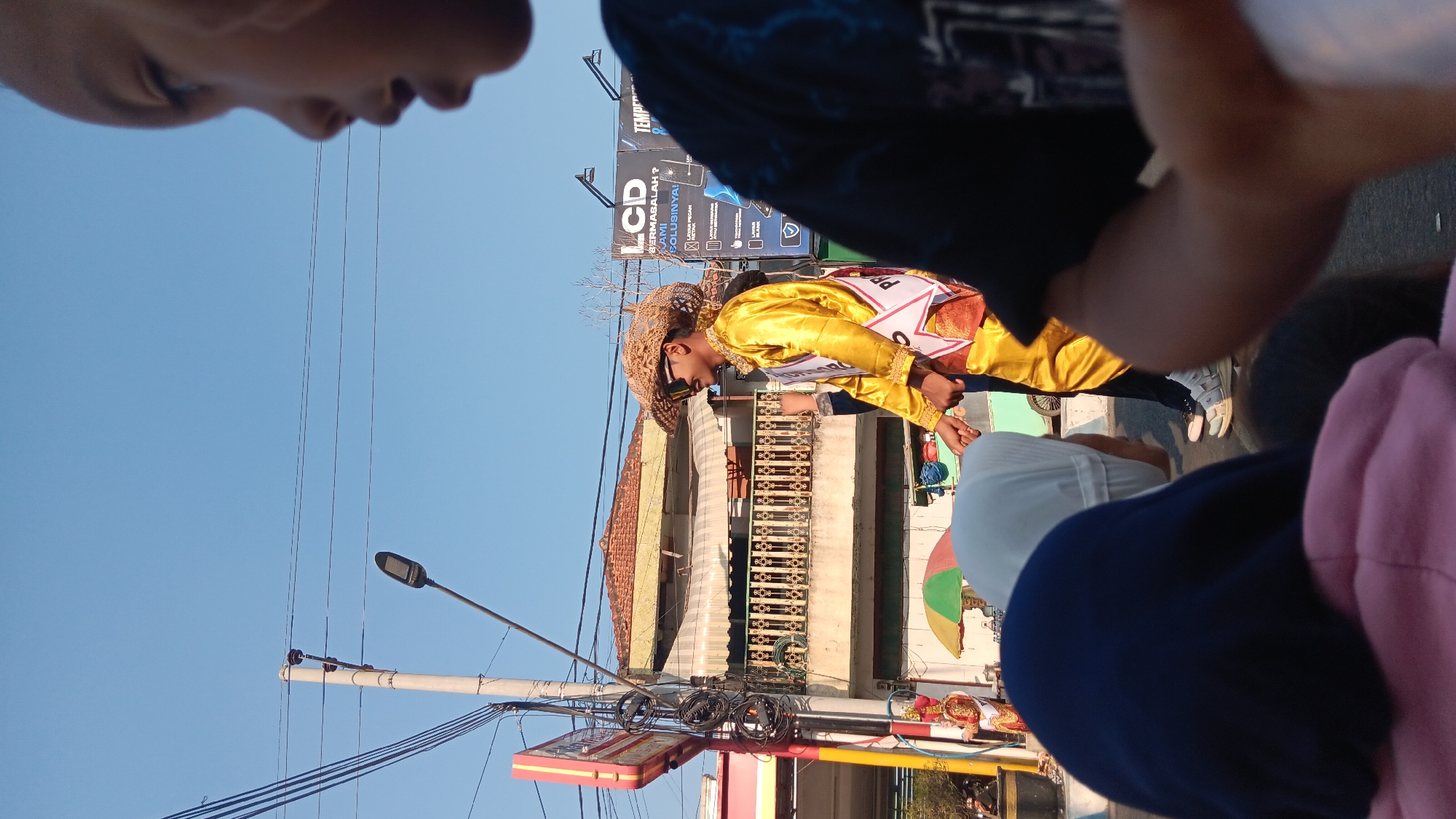 In Warujayeng, Indonesia, a young man, adorned in a vibrant gold satin outfit and a woven straw hat, participates in an outdoor event. He wears sunglasses and a sash that reads "PR". He is holding hands with another person, whose face is obscured. In the foreground, three individuals are visible from behind, with one wearing a dark blue hijab and another a pink hoodie, suggesting they are observing the event. The background features buildings with balconies and awnings, overhead power lines, a street lamp, and a large billboard advertising LCD solutions. The scene appears to be a daytime parade or festival, indicated by the costumes and the festive atmosphere.