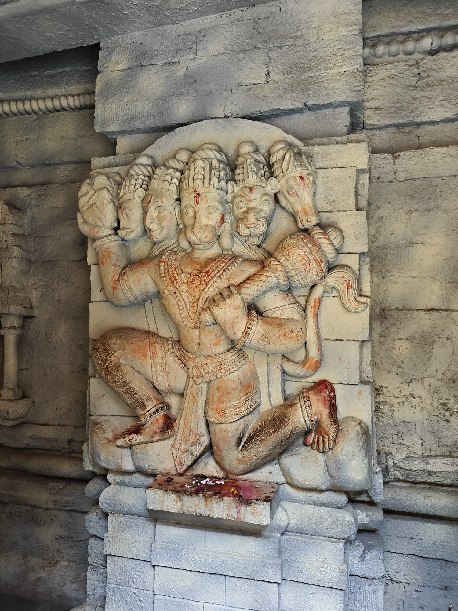 A detailed relief sculpture of a multi-headed deity, likely Panchamukhi Hanuman, is prominently featured on a textured wall within what appears to be a temple or religious structure in Manikaran, India. The sculpture depicts a powerful figure with five heads, the central one resembling a monkey, holding a mace (gada) with both hands. The deity is shown in a dynamic pose, with one knee bent and the other leg extended, resting on a base.
The sculpture is carved from a light-colored material, possibly stone or plaster, and shows signs of ritualistic worship. Reddish-orange pigment, likely kumkum or sindoor, is applied to the foreheads of each head, as well as on various parts of the body, including the arms, legs, and the mace. Darker reddish-brown stains, possibly from oil or other offerings, are visible on the deity's lower legs and the base of the sculpture. At the very bottom, small white flowers and additional red pigment are scattered, indicating recent offerings.
The surrounding walls are constructed from roughly textured, light-grey or off-white blocks, giving a sturdy and ancient feel to the setting. In the upper left background, a distinctive coiled or rope-like decorative element runs horizontally along the wall. The lighting in the scene is somewhat dim and indirect, suggesting an indoor or covered area. No discernible text is visible in the image. The overall scene conveys a sense of devotion and spiritual significance.