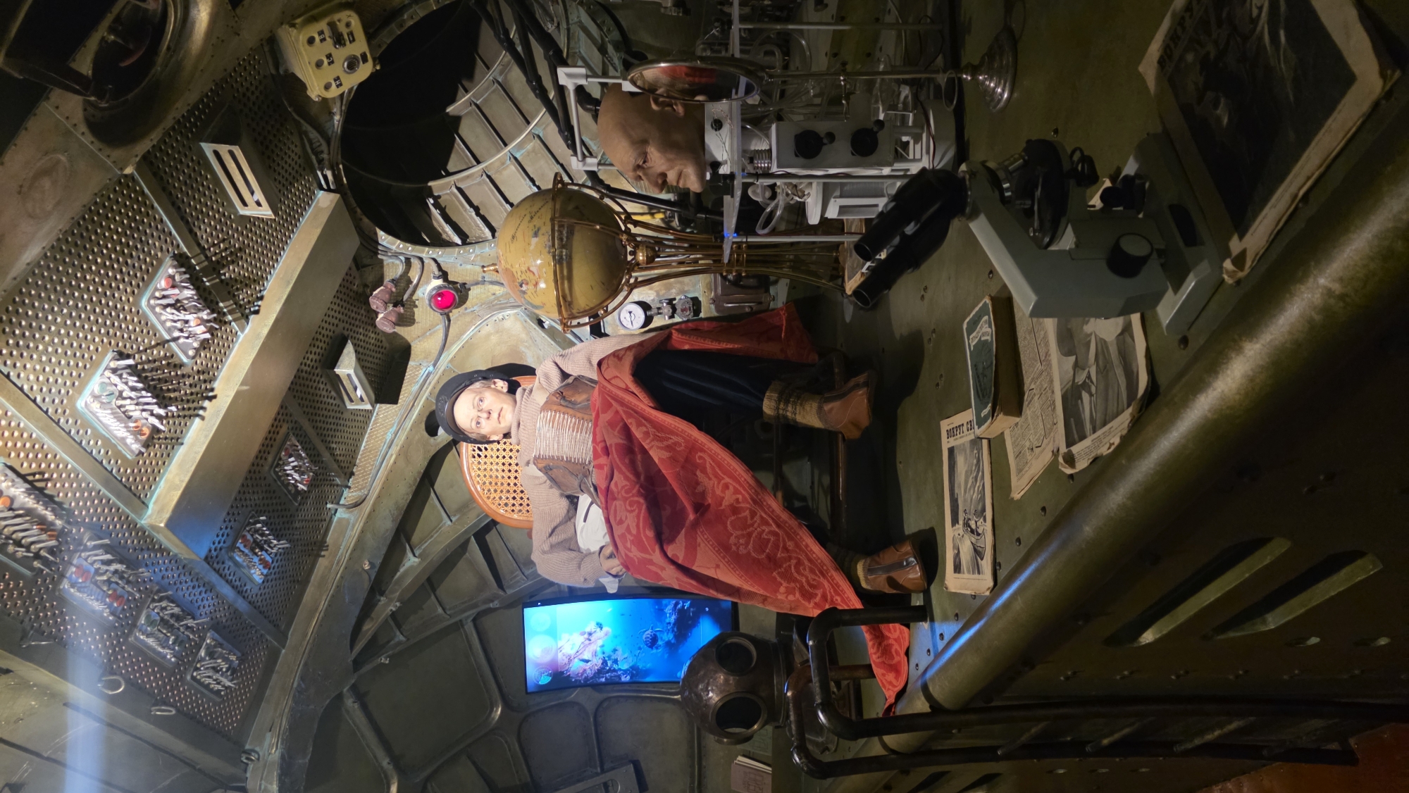 The image captures an elaborate, steampunk-inspired interior. The location is unknown. The central subject is a mannequin dressed in period clothing, seated in a chair. Surrounding the mannequin are various scientific instruments, including a globe, a microscope, and control panels filled with switches and gauges. A screen displays an underwater scene. Other details include a diving helmet, stacks of newspapers, and exposed pipes. The environment suggests a vintage submarine or laboratory.