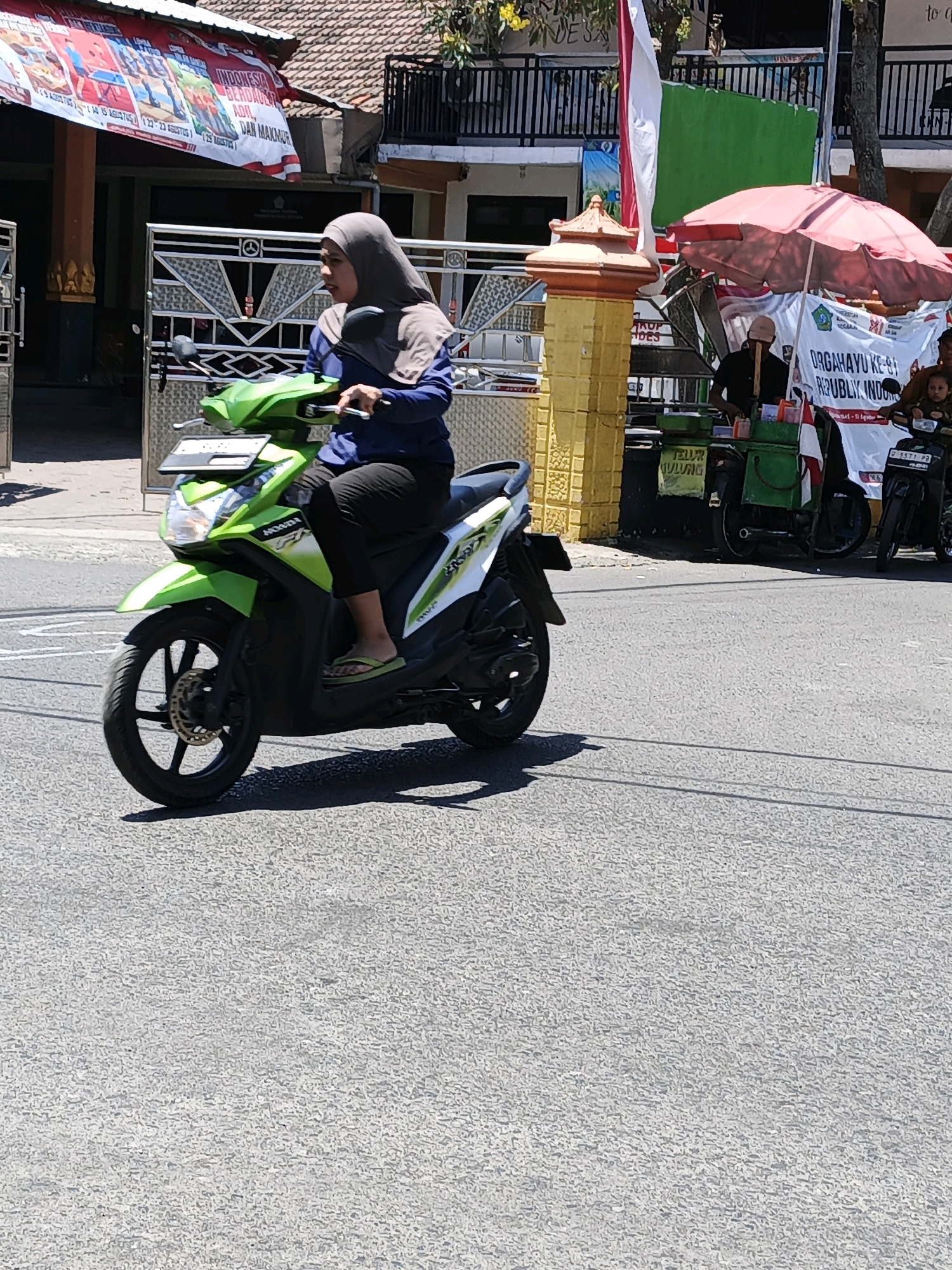 A woman wearing a hijab and a blue shirt rides a green Honda scooter on a paved road in Ketegan, Indonesia. She is wearing black pants and light-colored sandals. The scooter has a license plate and a front fender. In the background, there is a building with a red and white banner, a red umbrella covering a food stall, and another scooter with a child on the back. The scene is illuminated by sunlight, casting shadows on the road.