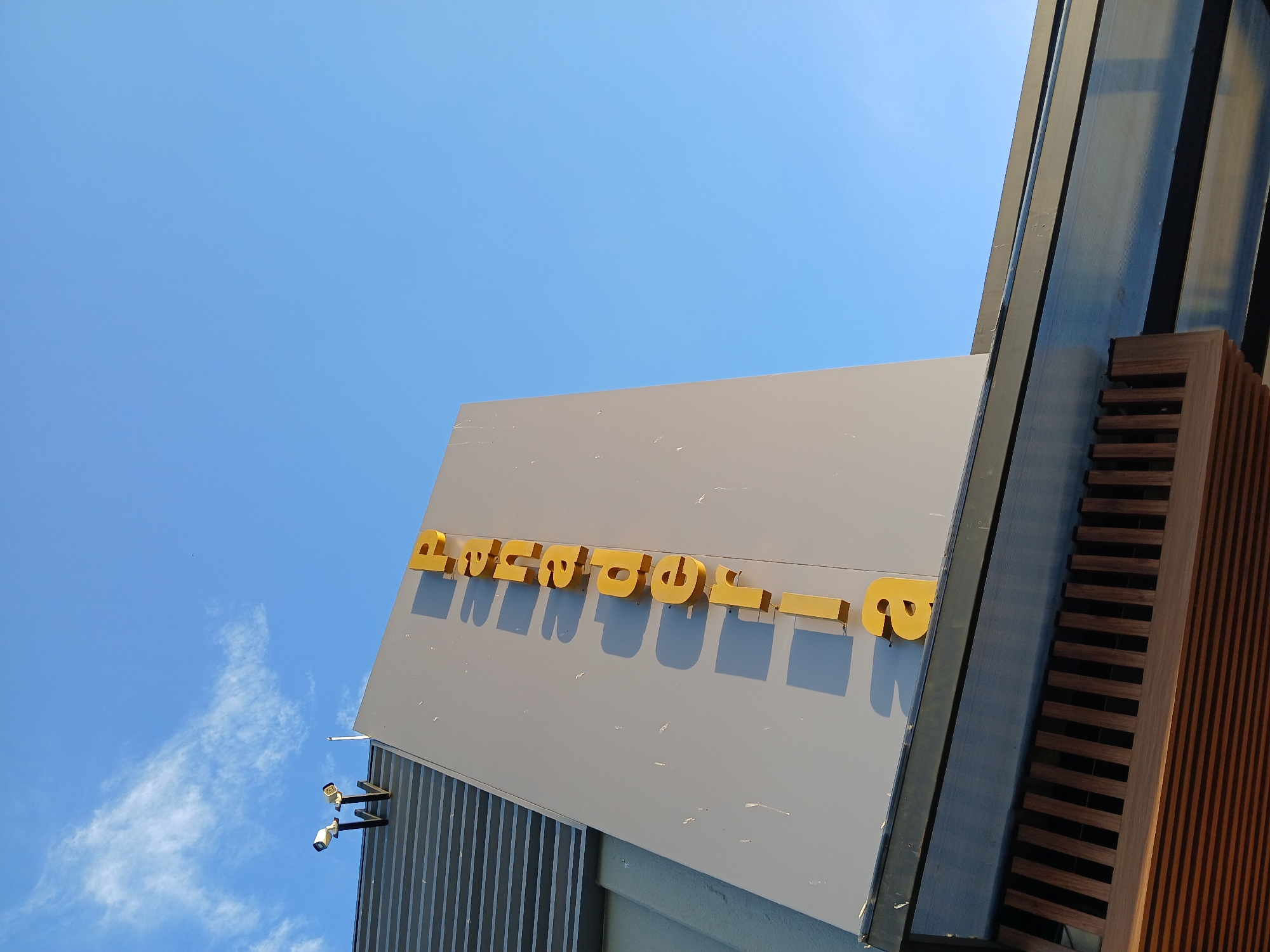 A low-angle shot shows a sign with the word "AVENTOURS" in yellow capital letters, mounted on a gray surface attached to a building. The upper right corner of the image contains vertical slats of wood and black metal. The building has visible security cameras. The sky is blue and clear. The location is Ciudad Guayana, Venezuela.