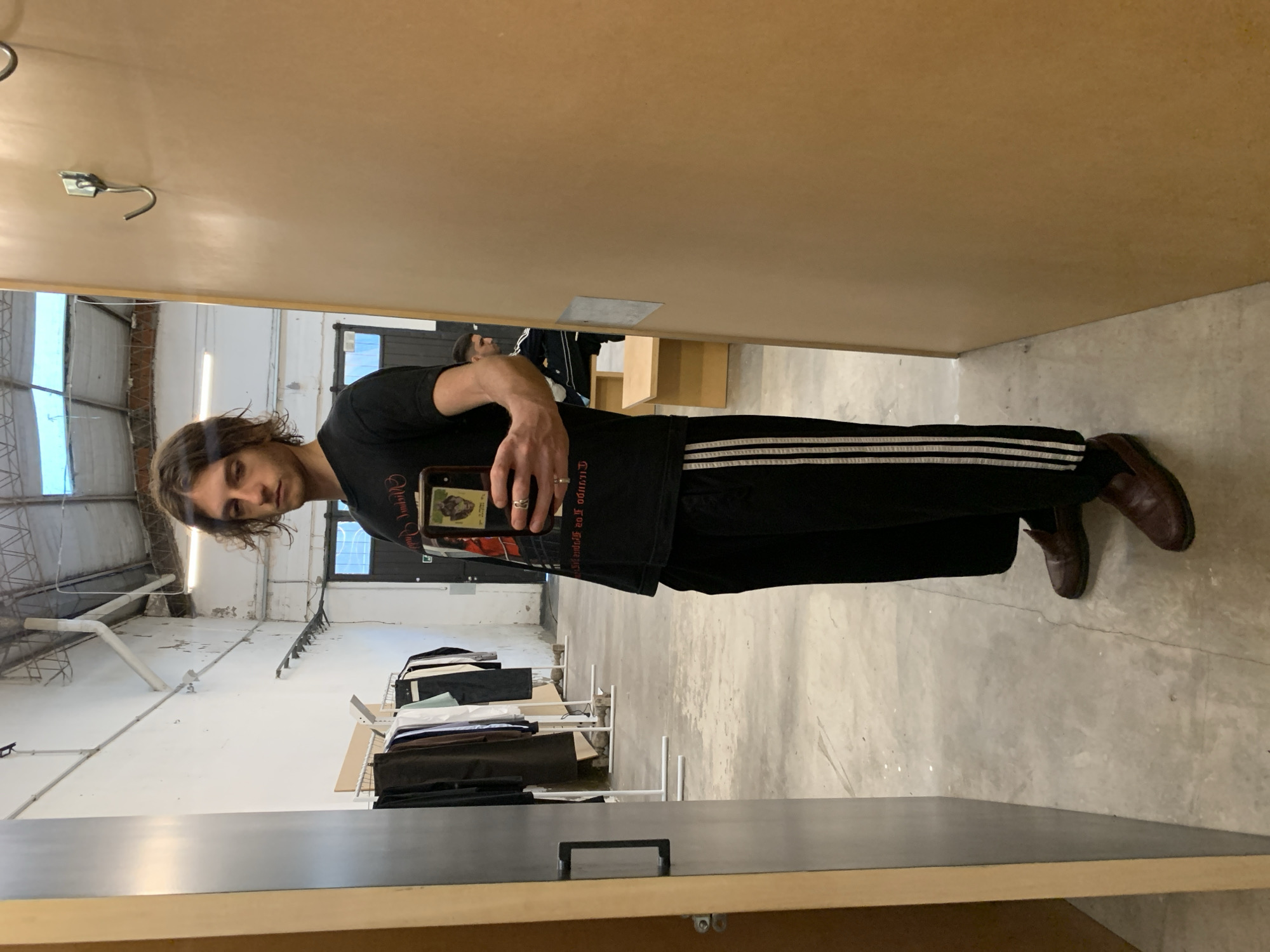 A young man takes a mirror selfie in what appears to be a changing room or designated mirror area within an industrial-style interior space, likely in Buenos Aires, Argentina. The image is captured from his reflection.
The man, with medium-length wavy brown hair, looks directly at the camera with a neutral expression. He is wearing a black short-sleeved t-shirt with light-colored, reversed text on the front that reads "EL CUERPO Y EL TIEMPO..." (The Body and Time...). He also wears black track pants with three white stripes down the sides and dark brown slip-on loafers. He holds a smartphone in his right hand, which has an image resembling an old illustration of a figure, possibly a dog or bear, on its screen or case. A silver ring is visible on his ring finger.
The background reflected in the mirror reveals a spacious, raw industrial setting. This includes high ceilings, exposed metal support structures, large windows or skylights letting in natural daylight, and concrete floors. Clothing racks, displaying a variety of folded and hanging garments, are visible behind the man, suggesting a retail store, showroom, or a pop-up market. The walls of the mirror enclosure are made of a light brown wooden or particle board material, with a silver hook visible near the top. The overall ambiance is modern and urban, fitting for a converted industrial space.