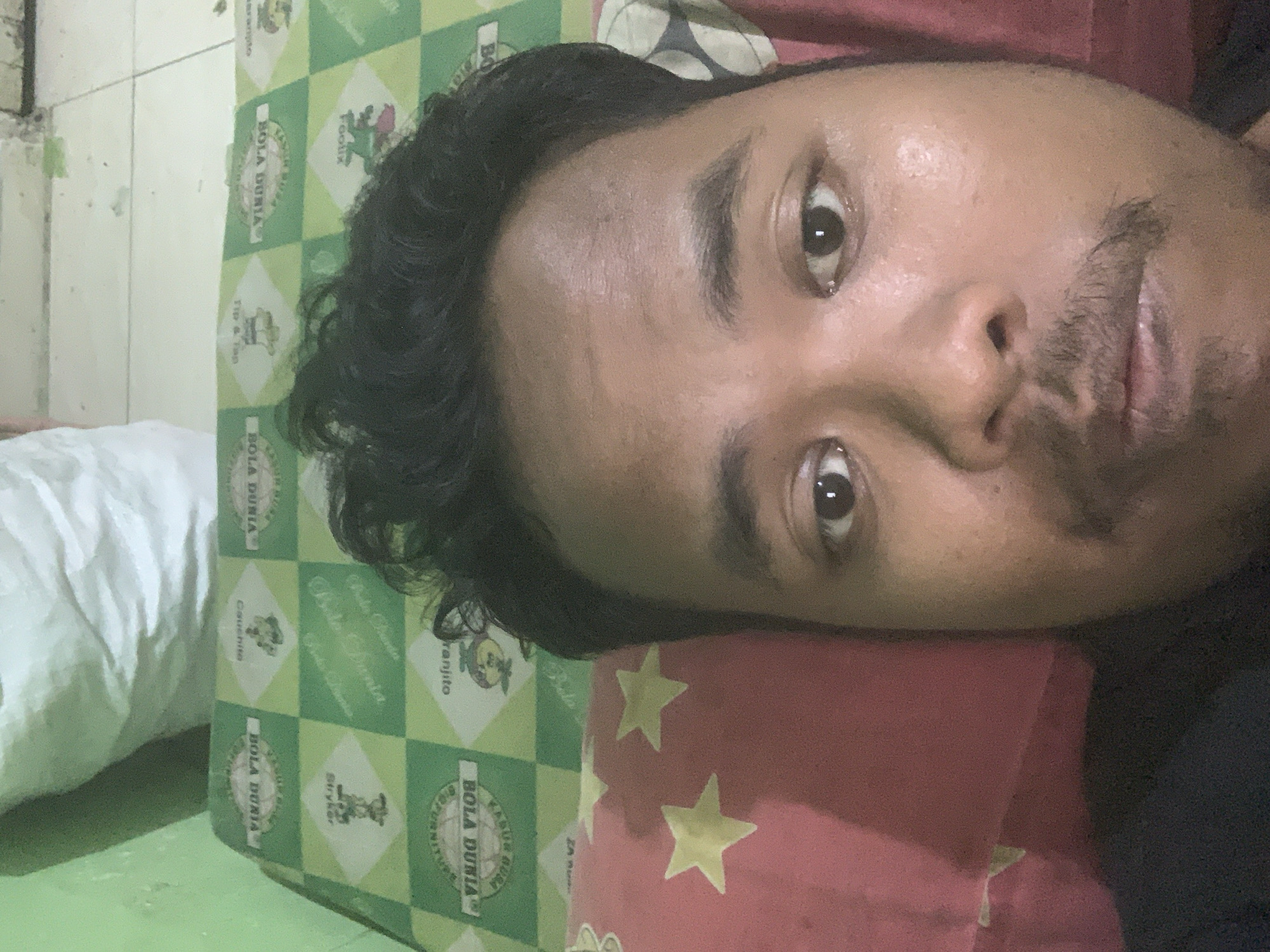 A close-up selfie shows a man's face as the primary subject. He has dark, curly hair and a mustache. He is lying down, with a red pillow with yellow stars visible beneath him on the left side of the frame. Behind him, a green and white checkered mattress is visible, featuring text that appears to be in Indonesian, including "BOLA DUNIA" and "Kasur". The background includes a light-colored wall and floor tiles, suggesting an indoor environment. The lighting is dim, and the image quality is somewhat grainy. The overall scene appears to be a casual, personal moment captured in a bedroom setting. No specific location in Palembang, Indonesia is identifiable from the image itself; it depicts a common domestic setting.