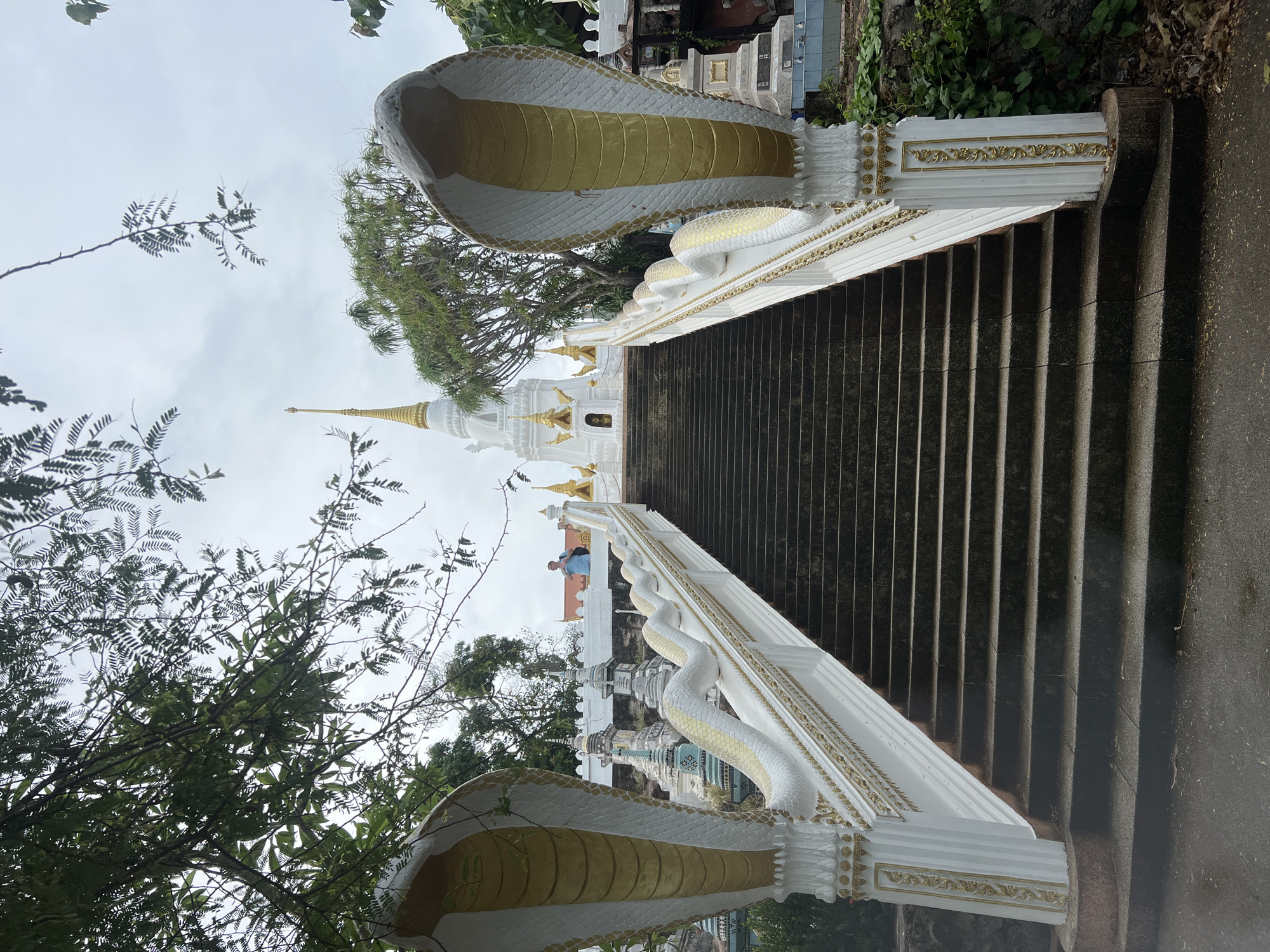 The image depicts a staircase leading to a white and gold temple in Baan Hua Thanon, Thailand. The staircase is flanked by white serpent sculptures. The environment includes trees and other greenery. A person is visible near the temple.