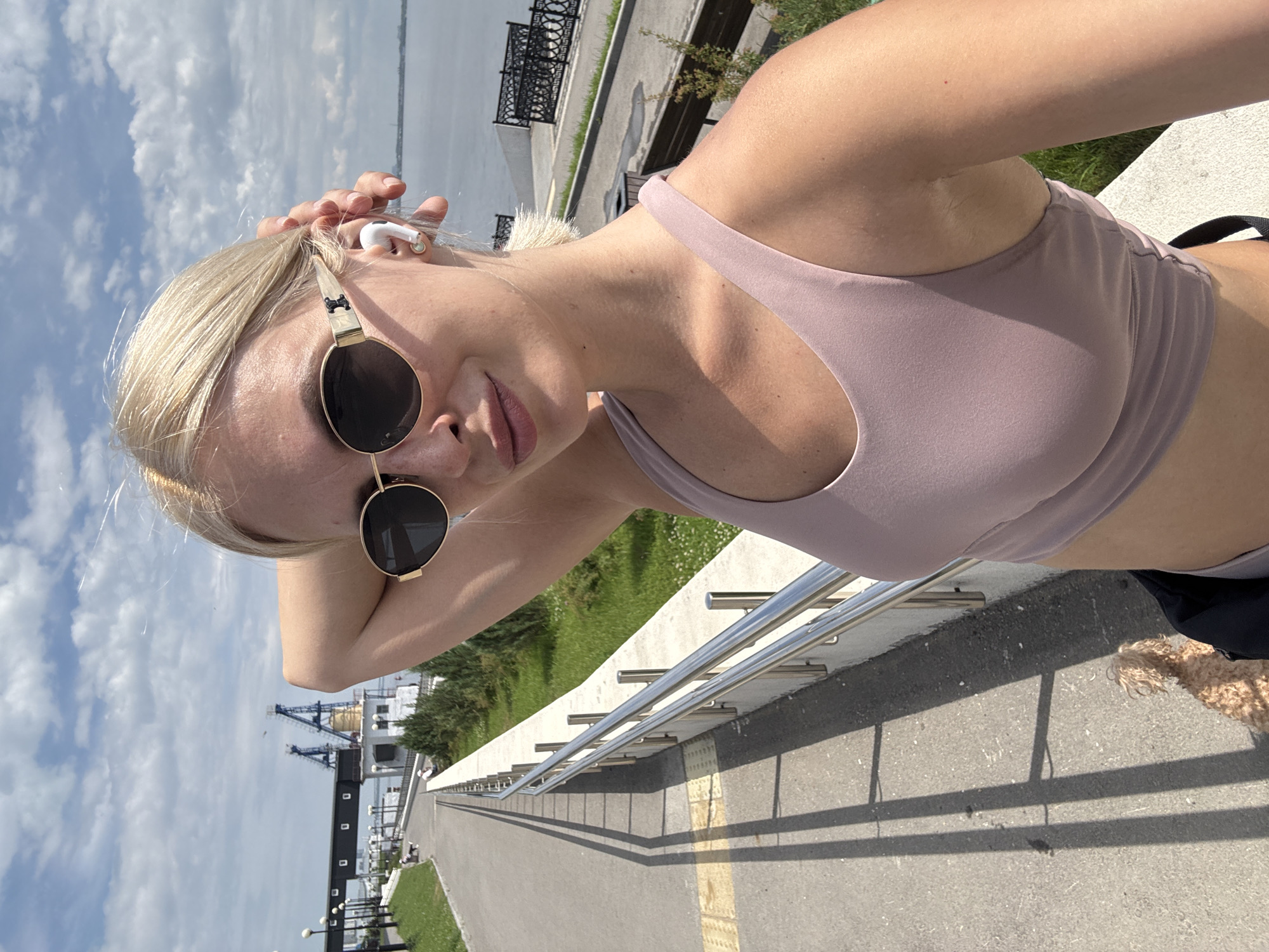 The image shows a woman taking a selfie on a sunny day in Saratov, Russia. She is wearing sunglasses and a light pink sports bra, with a white wireless earbud in her right ear. Her left hand is touching her hair. In the background, a paved walkway runs alongside a grassy area with trees and a metal railing. Further in the background, there is a body of water, industrial cranes, and a cloudy sky. A small dog is visible on the walkway in the lower left of the frame. The scene appears to be a promenade or park area near a waterfront.