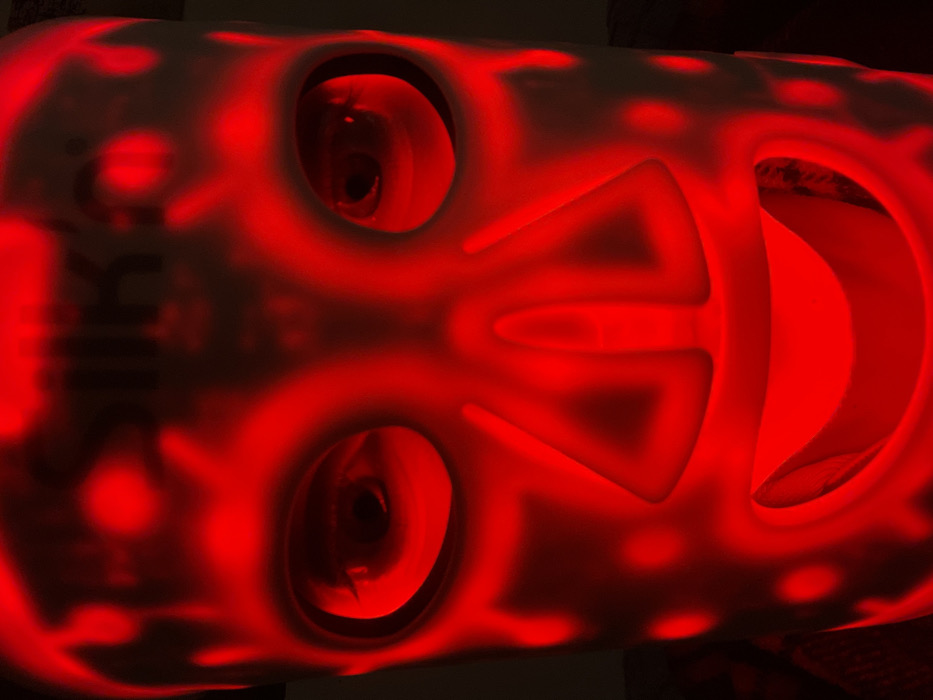 A frontal, close-up photograph depicts an individual wearing an illuminated facial mask, located in Monsummano Terme, Italy. The mask is a light-colored, rigid device covering the entirety of the face, featuring apertures for the eyes and mouth. The surface of the mask is illuminated with intense red light, emanating from patterned dots, triangles, and rounded shapes distributed across the forehead, cheeks, nose bridge, and chin. The brand name "SILK'N®" is faintly visible in dark text on the upper forehead area of the mask. The subject's eyes are discernible through the mask's eye openings, staring directly forward, with pupils exhibiting slight dilation. The ambient environment is dimly lit, enhancing the prominence of the mask's red luminescence.