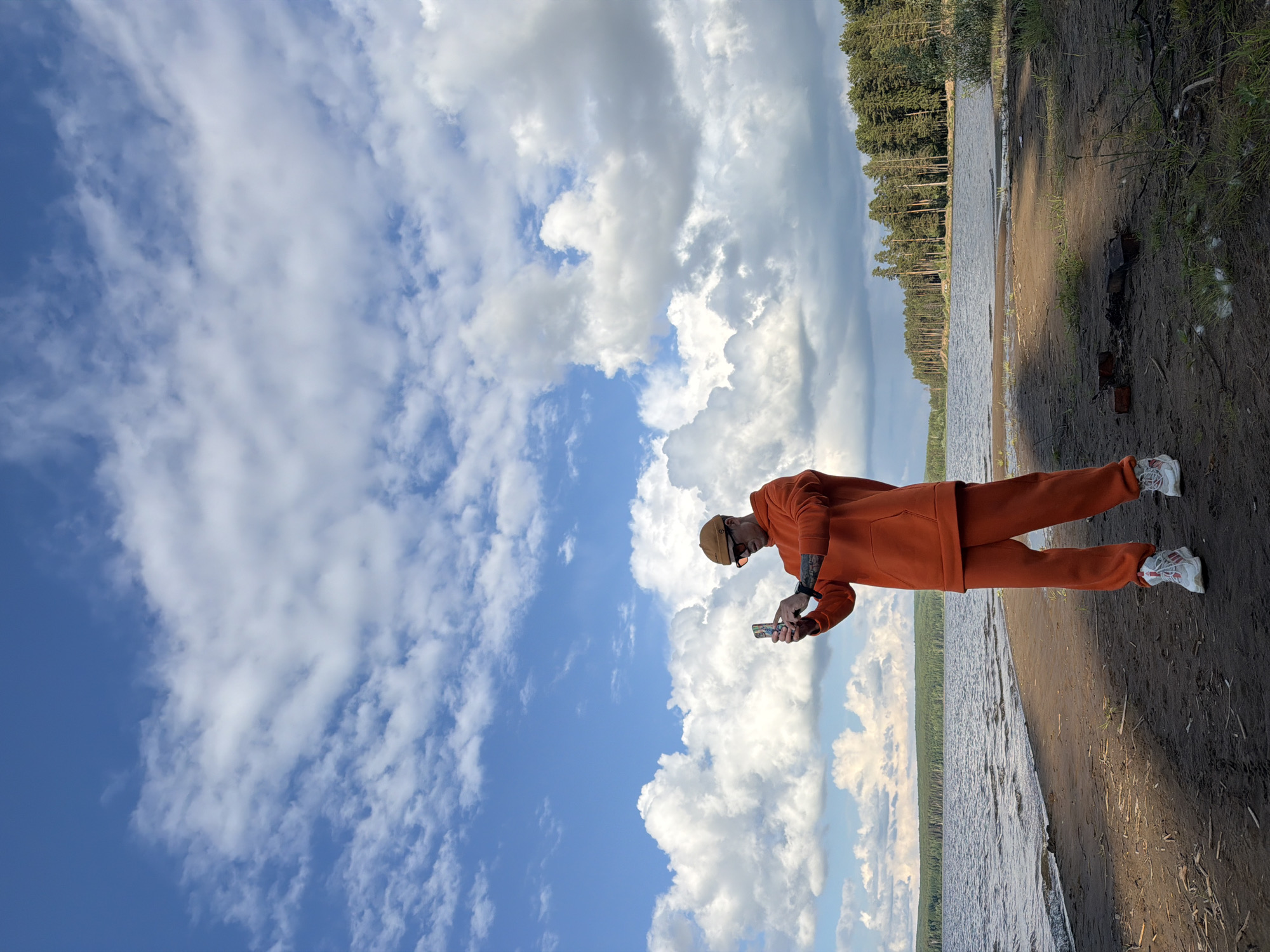 An outdoor, full-body shot captures a person standing on a muddy and sandy shoreline next to a body of water, likely a lake or wide river, in Chaikovsky, Russia. The individual, appearing male, is wearing an orange tracksuit (hoodie and pants), a light brown beanie, sunglasses, and white sneakers with red accents. Tattoos are visible on their forearms and wrists. They are looking down intently at a smartphone held in both hands, possibly texting, browsing, or capturing an image. A watch is visible on their left wrist.
The water surface is slightly choppy, reflecting the overcast sky. Across the water, a dense line of coniferous trees forms a distant forest. The immediate foreground shows the damp, dark earth of the shore, with some small pieces of wood or logs scattered near the person's feet, suggesting a past campfire. The sky above is largely covered with large, fluffy white and grey clouds, with patches of bright blue visible. The overall lighting suggests it is daytime, possibly late afternoon. No discernible text is visible on the person's attire or phone screen. The scene depicts a serene natural environment.