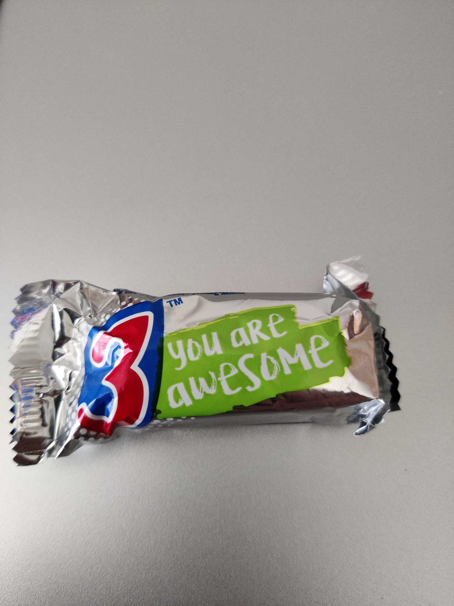 A close-up, eye-level shot shows a chocolate bar wrapper with the phrase "You Are Awesome" printed in white on a lime green background. The wrapper is mostly silver and crinkled, with a large blue and red numeral "3" on the left side. The background is a plain, light gray surface, possibly a table or desk. The location of this image is Lower Paxton Township, United States of America.