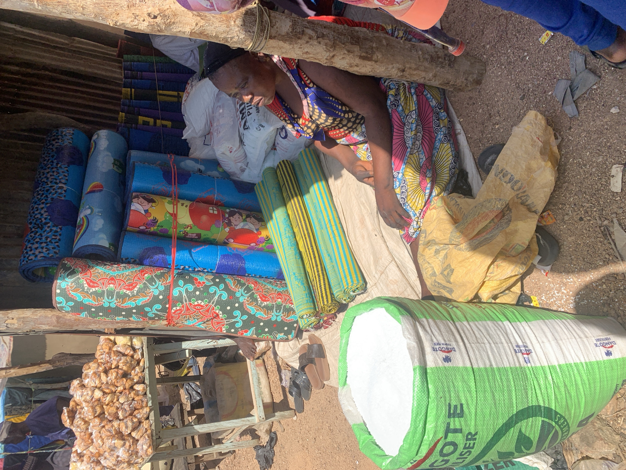 The image depicts an outdoor market stall in Nigeria. The primary subjects are a woman, likely a vendor, and various goods for sale. The woman is wearing a brightly colored, patterned dress and appears to be resting or attending to her merchandise.
The objects for sale include an assortment of rolled-up mats, some with colorful patterns and cartoon characters, others with striped designs. There are also several large sacks, one of which is predominantly green and white and contains a white granular substance, possibly fertilizer given the visible text on it. Other sacks are a pale yellow color and appear to be made of woven material. A pile of small, brown, individually packaged food items, possibly snacks or sweets, is also visible, neatly arranged on a shelf. A pair of sandals lies on the ground near the woman. The setting is rustic, with corrugated metal sheeting visible in the background and loose gravel or dirt on the ground. A wooden beam runs overhead, forming part of the stall's structure.
The time of day appears to be daytime, with natural light illuminating the scene. The weather seems to be warm, typical of an outdoor market in Nigeria. The overall impression is one of an active, albeit somewhat cluttered, marketplace.
Visible text on the green and white sack reads: "DANGOTE FERTILIZER" and "ER" at the bottom edge. The phrase "DANGOTE FERTILISER" is repeated in smaller print along the top band of the sack.