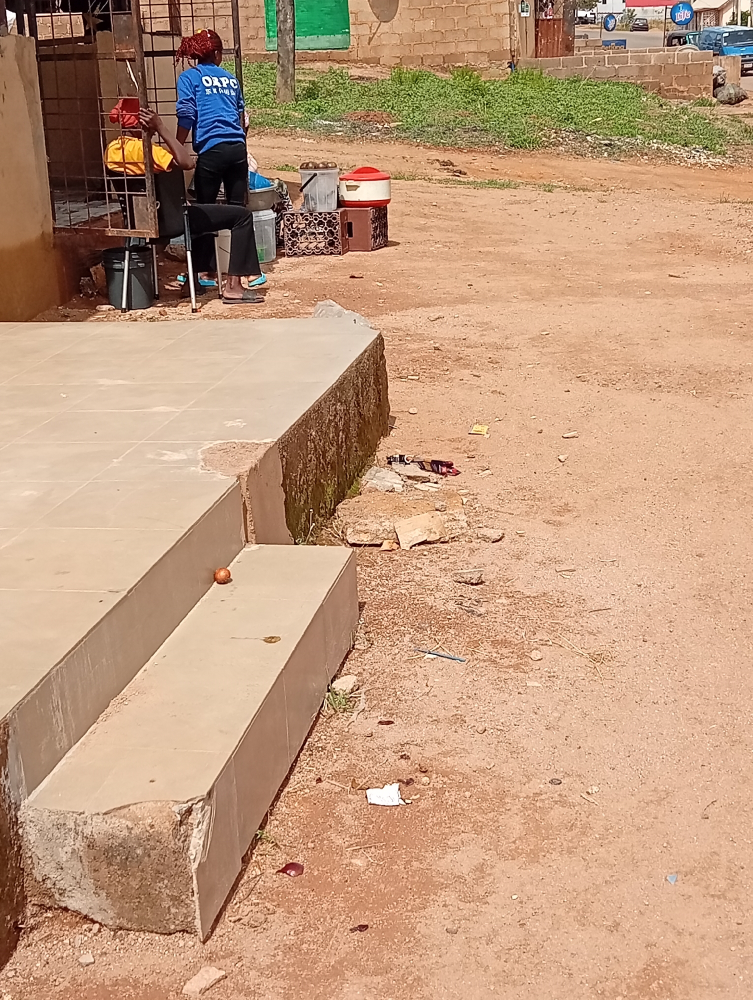 The image shows a scene in Jos, Nigeria. In the foreground, there are light brown tiled steps with an orange object (possibly an onion or tangerine) on the lower step. The ground is reddish-brown soil with scattered debris and vegetation. In the middle ground, two people are visible near a building with a barred opening. One person is wearing a blue shirt with the letters "ORPC" on the back and standing. The other person is seated in a chair, partially obscured by the bars. Behind them are various containers and objects, including a red and white cooler and plastic crates. In the background, there is a green area and a building made of brown bricks, with vehicles visible in the distance. A road sign is barely noticeable in the upper background.