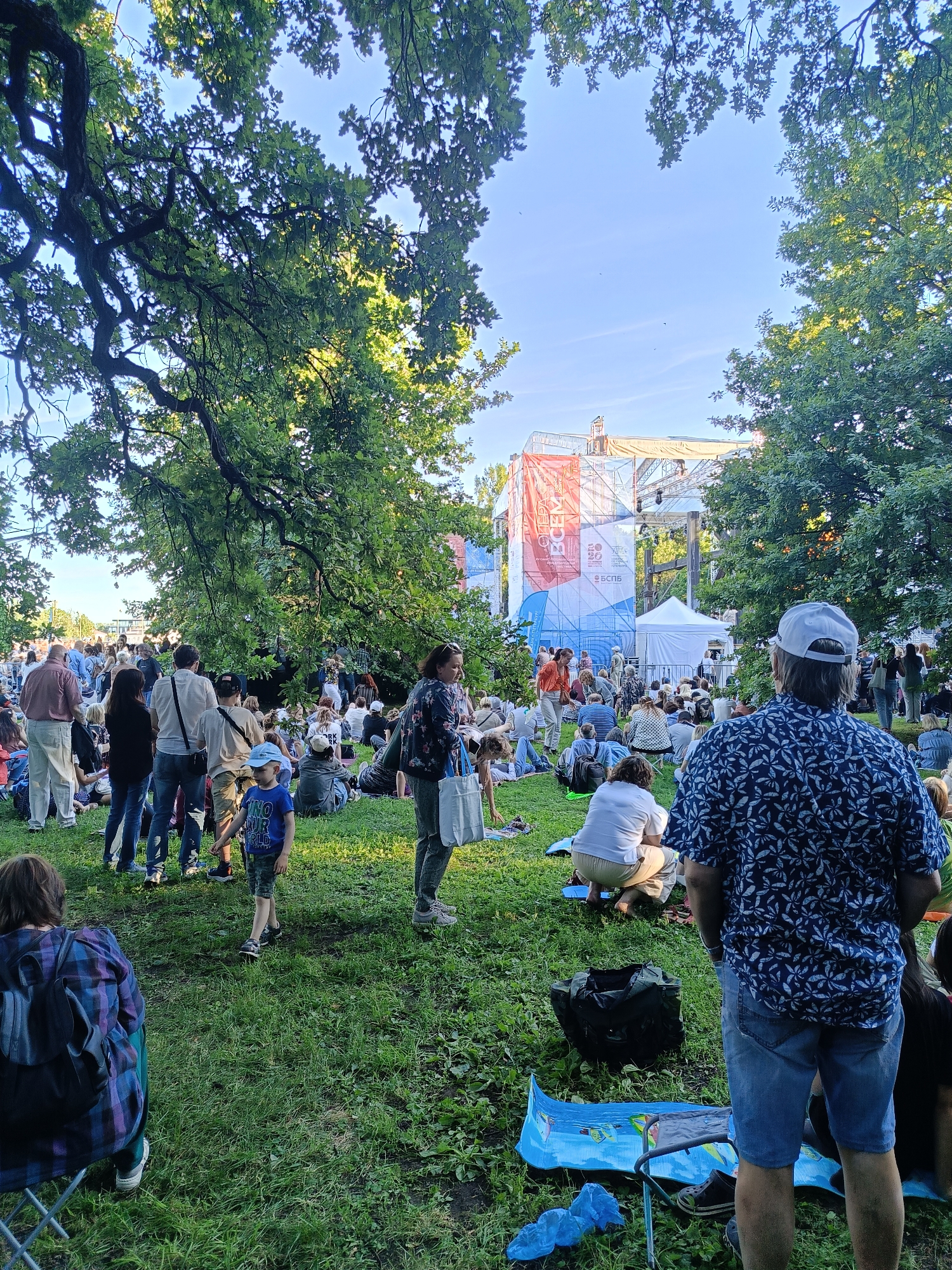 A crowd of people are gathered on a grassy area in front of a large outdoor stage structure. The stage is covered with banners and scaffolding, and appears to be set up for an event. The people are sitting and standing on the grass, some are picnicking, and others are watching the stage. The environment is a park, with large trees providing shade. The sky is clear and blue. The time of day appears to be late afternoon or early evening, as the sun is casting long shadows. The overall atmosphere is relaxed and festive.