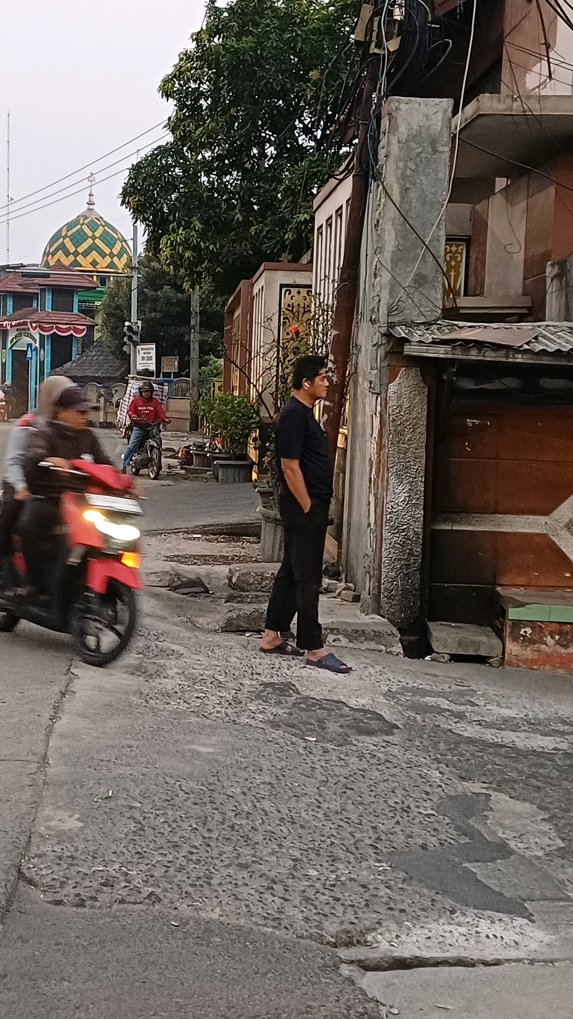 The image captures a street scene in Cibinong, Indonesia, during what appears to be late afternoon or early evening. A man in black attire, wearing blue sandals, stands with his hands in his pockets, facing a building on the right. He has short, dark hair and is wearing earphones. Behind him, a red motorcycle with two riders is in motion, the front rider wearing a white hijab and a dark jacket, and the rear rider wearing a dark jacket and a light-colored headscarf. Further in the background, another person rides a motorcycle, wearing a red shirt and a helmet.
The environment features a paved road with visible cracks and uneven surfaces. To the left, a mosque with a green and yellow checkered dome is visible, along with other buildings. Trees line the street, and electrical wires are strung overhead. The building the man is facing has a textured concrete pillar and a weathered wooden door or shutter. The lighting suggests a diffused, possibly overcast sky.