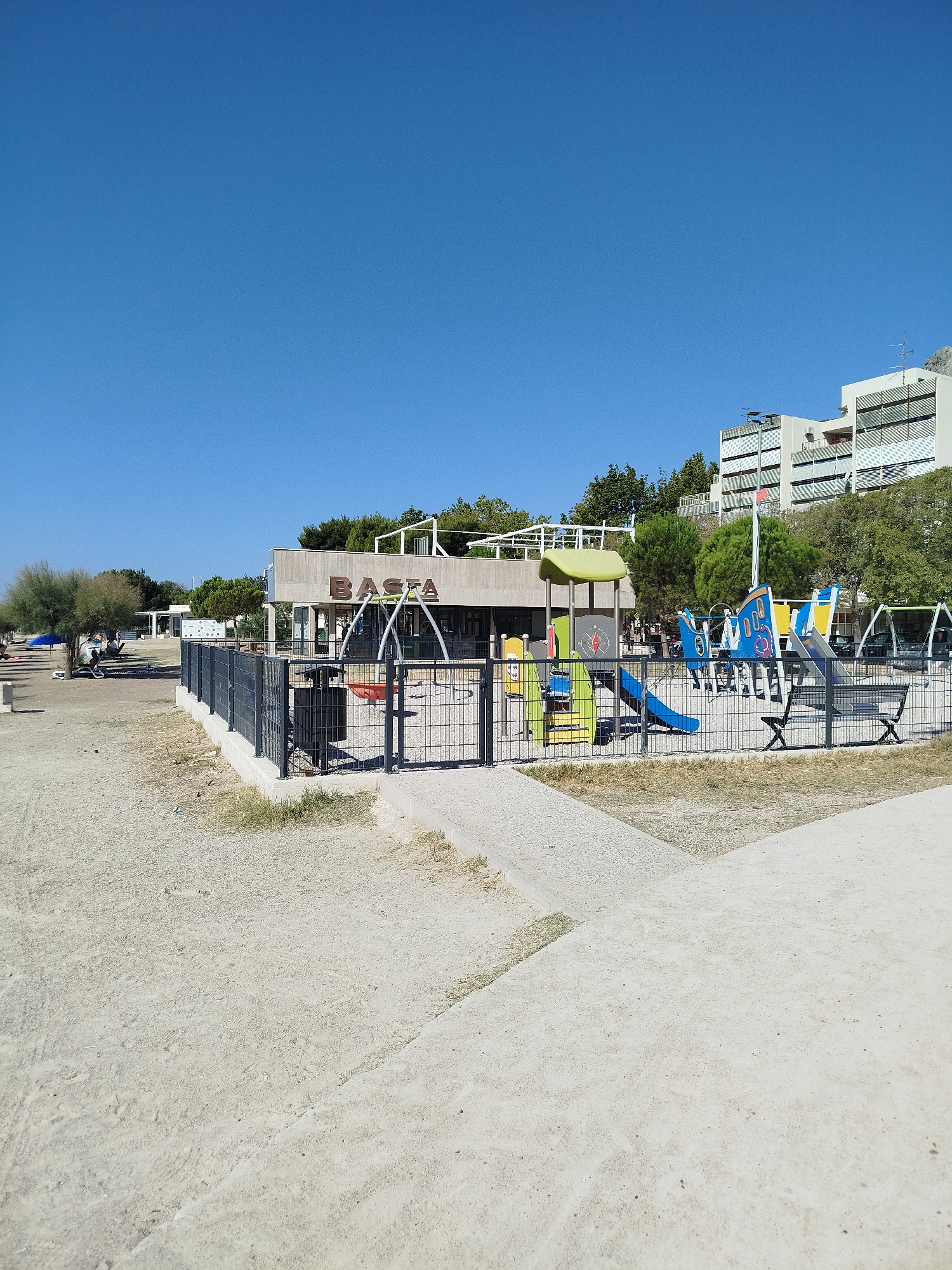 The image depicts a children's playground and a cafe in Omiš, Croatia, on a clear, sunny day. The playground features a multi-colored climbing structure with slides, a ship-themed play area, and swings, all enclosed by a black metal fence. Adjacent to the playground is a cafe with a sign reading "BASTA". The area surrounding the playground and cafe consists of sand and gravel. In the background, there are trees, a building with balconies, and a glimpse of a rocky hillside. The scene is devoid of people actively using the playground or cafe.