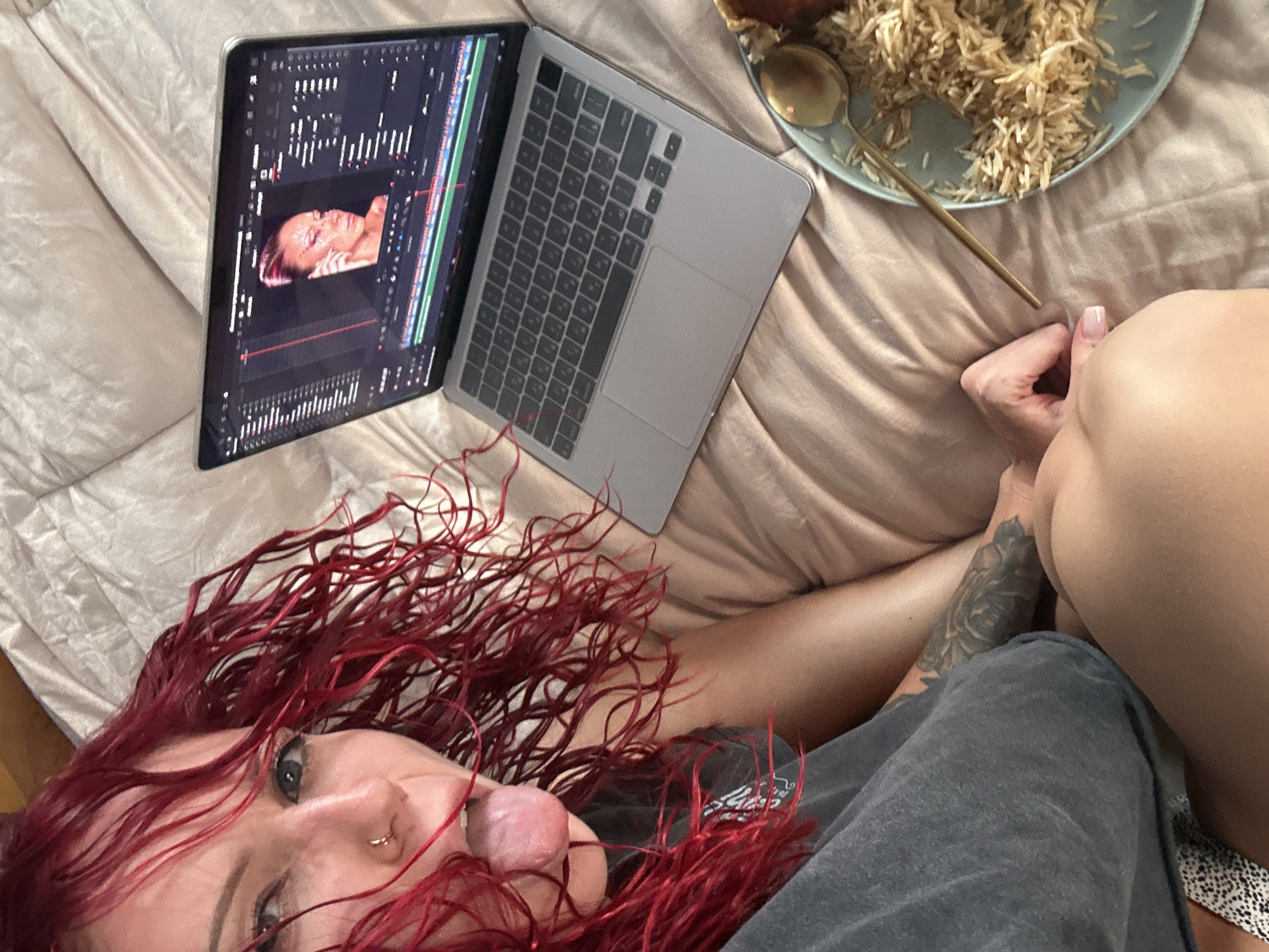 The image depicts a person with red curly hair lying on a bed in a supine position. The person has fair skin, facial piercings, and is wearing a grey garment. Their tongue is extended. The bed is covered with a light-colored, possibly beige, sheet. A laptop is positioned on the bed, open and displaying editing software with a female face visible within the program. A plate containing rice is also visible on the bed, with a golden spoon next to it. A partially exposed tattoo is visible on the person’s arm. The location is presumed to be an indoor setting and does not provide enough information to determine if the location is in North Kuta, Indonesia.