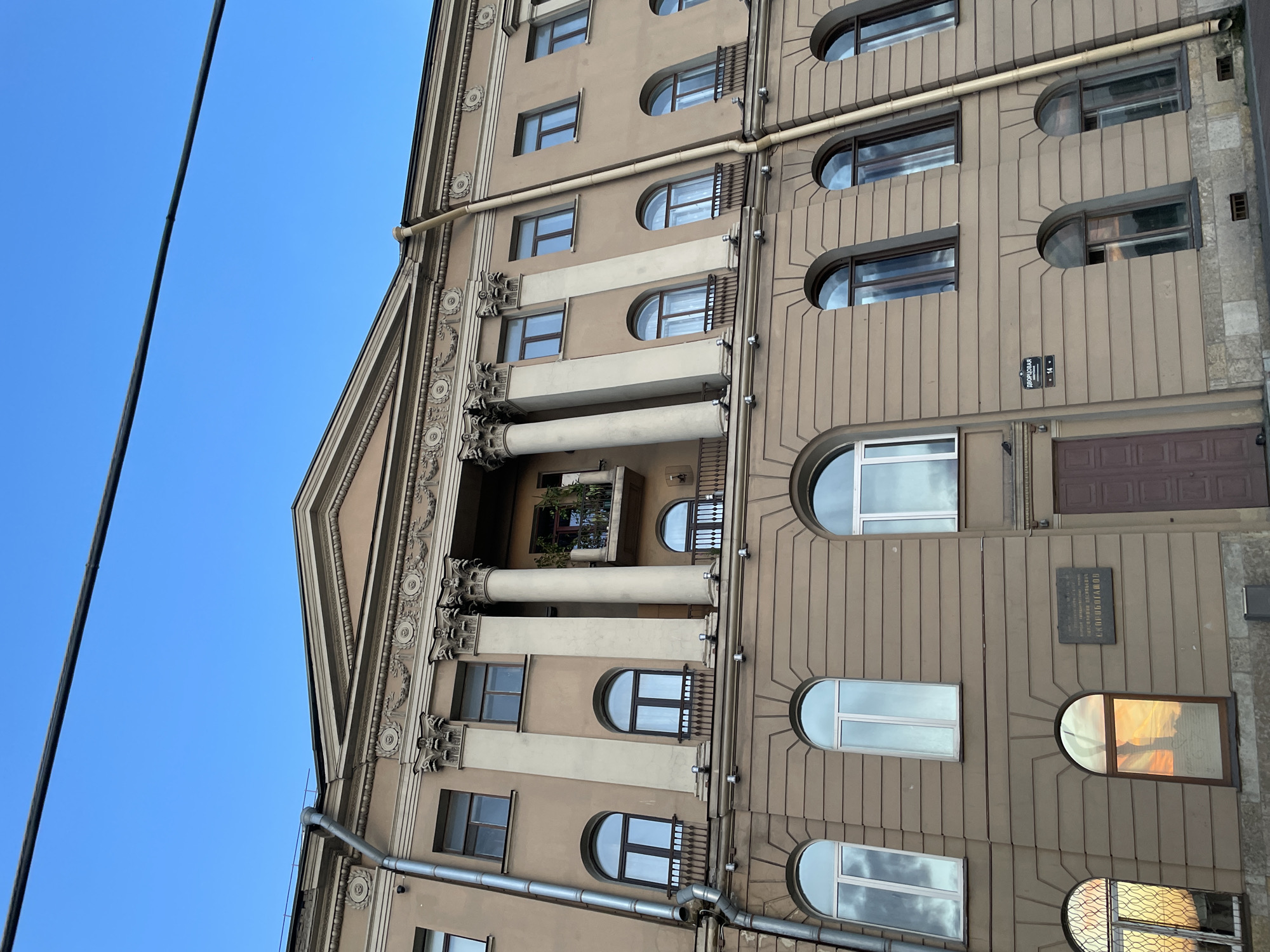 A low-angle, eye-level exterior shot captures the facade of a multi-story building under a clear blue sky. A black wire stretches diagonally across the top of the frame. The building is light brown with a classical architectural style. The upper facade features a triangular pediment adorned with decorative molding. Below the pediment, a row of tall, fluted Corinthian columns supports a balcony with an ornate railing. Several windows punctuate the upper level, with arched windows featuring small balconies on the lower level. The middle level of the building has a series of arched windows with decorative ironwork. Below these, another row of rectangular windows is visible. A metal downspout runs vertically along the left side of the building. A dark brown door is centered on the ground floor, flanked by windows. A rectangular plaque with text is mounted on the wall to the left of the door. The reflection of a sunset sky is visible in some of the lower windows.