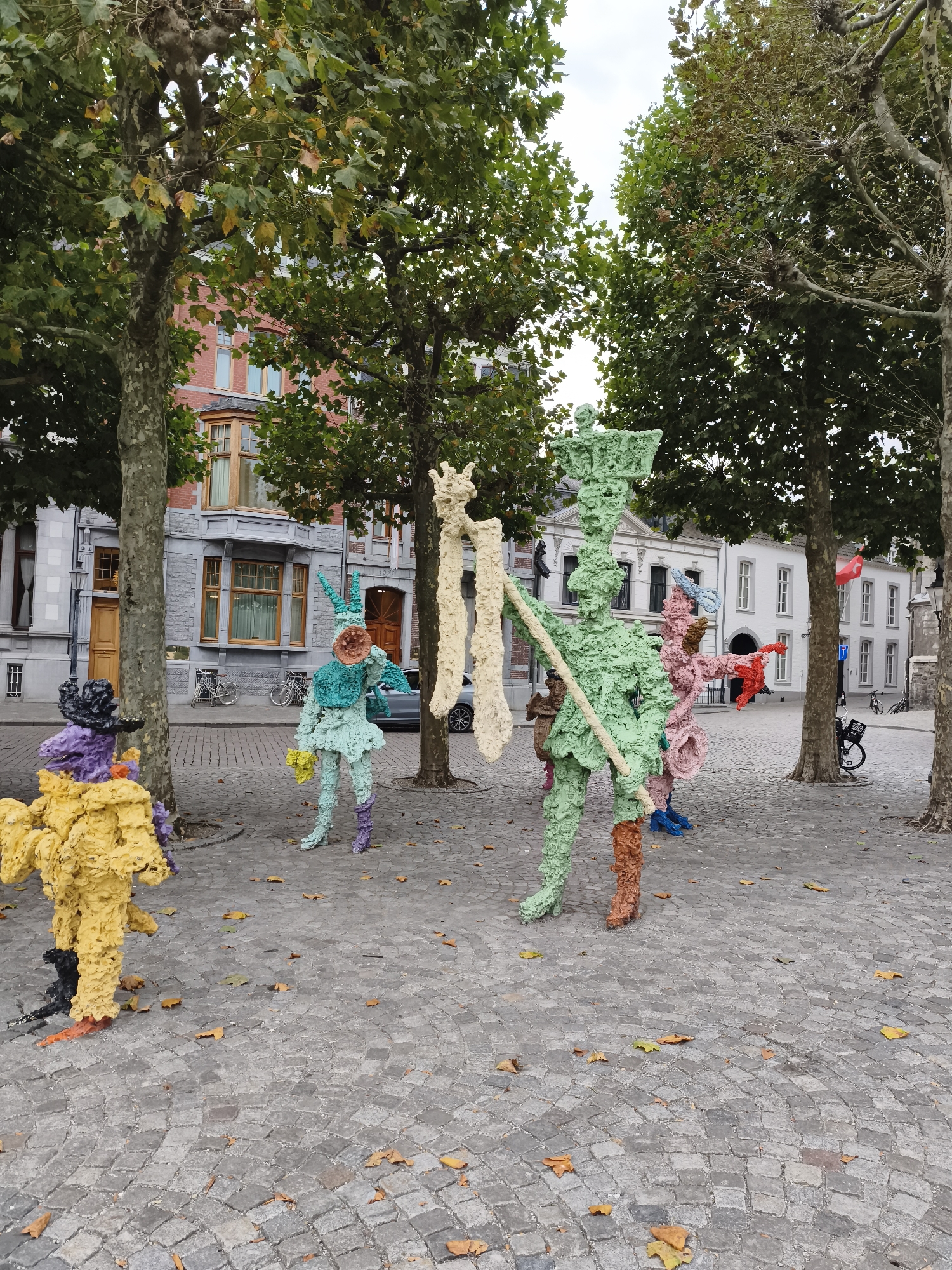 The image depicts an outdoor public art installation in the city of Maastricht, Netherlands. The installation consists of several large, colorful, textured sculptures resembling humanoid figures or creatures. The sculptures are predominantly made of a coarse, porous material, painted in bright hues such as yellow, purple, teal, green, pink, and light beige.
In the foreground, a yellow sculpture is positioned on the left, with purple and black elements at its base. To its right stands a teal sculpture with a distinctive pointed headpiece and a circular detail on its chest. This sculpture holds a yellow object. Further to the right, a tall green sculpture stands prominently, holding a light beige, rod-like object and a beige strip. To its right is a pink sculpture with red accents.
The sculptures are situated on a cobblestone pavement, scattered with fallen autumn leaves. Large trees with dense foliage, some showing signs of autumn colors, provide a canopy over the scene. In the background, buildings with red brick and grey stone facades are visible, suggesting an urban setting. One building has a prominent bay window with wooden framing. In the far background, white buildings with multiple windows and a Dutch flag flying from a pole can be seen. Bicycles are parked near one of the buildings. A modern car is partially visible on the right side of the image. The overall scene is set on a cloudy day.