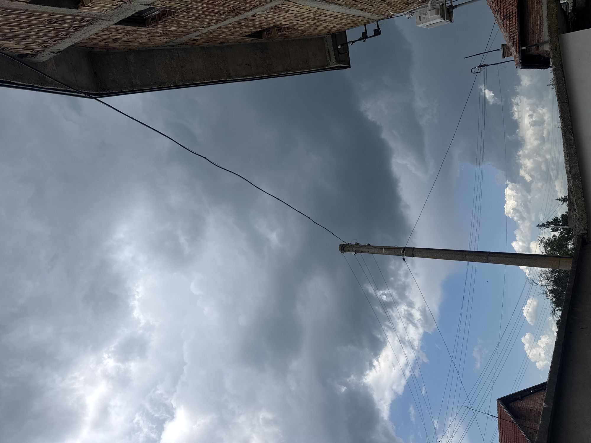 An upward-facing view shows a cloudy sky, portions of buildings, and utility poles and wires. The left side of the frame is occupied by a section of a brick building under construction. Utility wires run from the top-left corner diagonally across the sky towards a utility pole. The right side of the frame shows another building, a roof with red tiles, and some trees. The sky is a mix of dark gray storm clouds and patches of lighter clouds with blue sky visible. The location is Sarnevo, Bulgaria.