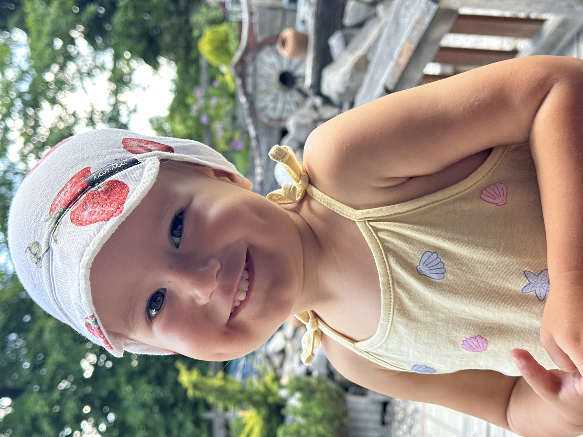 A young Caucasian child with fair skin, dark hair, and blue eyes is looking at the camera and smiling. The child is wearing a light yellow tank top with a pattern of seashells and a white hat with a strawberry pattern. The child is outdoors, with blurred green trees and a wooden structure in the background. The setting appears to be a garden or patio area. The child's facial features include dimples and visible teeth. The overall impression is of a happy and relaxed child in a natural environment.