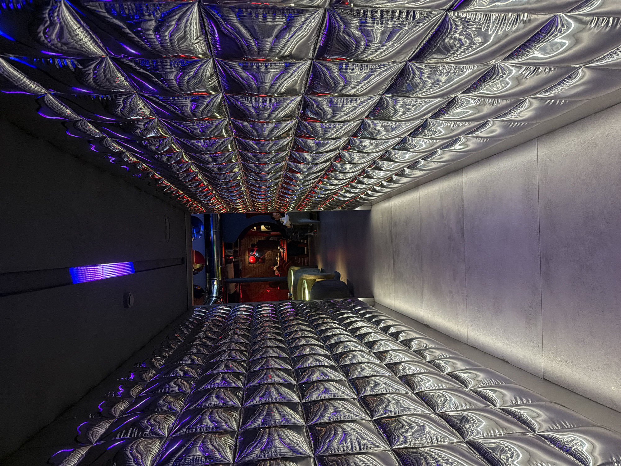 The image depicts a hallway with a unique design, possibly located in Moscow, Russia. The walls are painted a light gray color, with one side appearing darker than the other, likely due to lighting. The floor and ceiling are covered in a repeating pattern of reflective, quilted or padded squares, creating a futuristic or unconventional aesthetic. The lighting is varied, with hints of blue and red accents, suggesting the presence of colored light sources. In the distance, the hallway opens into a space that appears to be part of an interior with what looks like equipment or machinery. There is no evidence of people in the immediate view of the hallway. The floor and ceiling appear to be identical in material and pattern, suggesting an intentional design choice to create a mirrored or symmetrical effect.