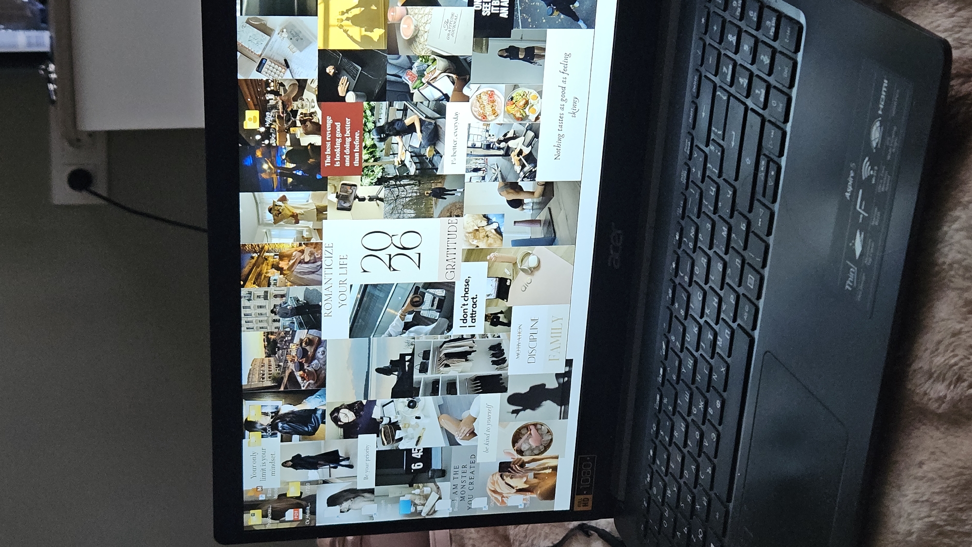 This is a close-up, eye-level, indoor shot of an open laptop displaying a digital vision board. The vision board is a collage of various images and text, including inspirational quotes, lifestyle photos, and personal goals. Some of the visible text includes "ROMANTICIZE YOUR LIFE," "20 26," "GRATITUDE," "The best revenge is looking good and doing better than before," "I don't chase, I attract," "MOTIVATION," "DISCIPLINE," and "FAMILY." The background behind the laptop is blurred and shows a wall with electrical outlets and a portion of a white surface, likely furniture. The laptop itself is a black Acer Aspire 5. The keyboard is visible in the foreground, and it appears to have Cyrillic and Latin characters on the keys. The lighting is dim, suggesting the room is not brightly lit.