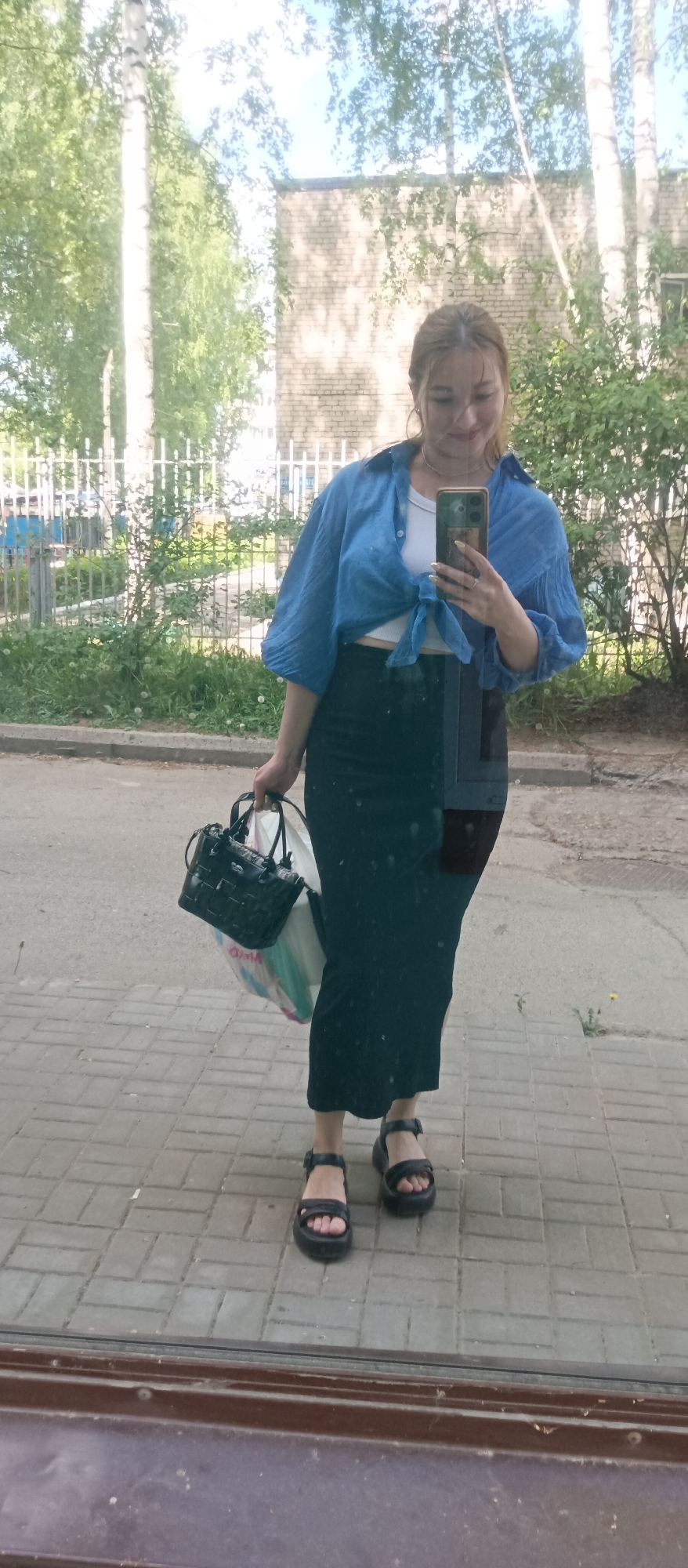 A woman stands on a brick sidewalk in an outdoor urban environment. She is holding a smartphone in her right hand, as if taking a selfie. She is wearing a blue, long-sleeved button-up shirt tied at the waist over a white top, a long dark skirt, and black platform sandals. In her left hand, she holds a black purse and a white shopping bag. Behind her, there is a building, trees, and a white fence. The scene appears to be outdoors on a sunny day. The city is identified as Unknown, Unknown.