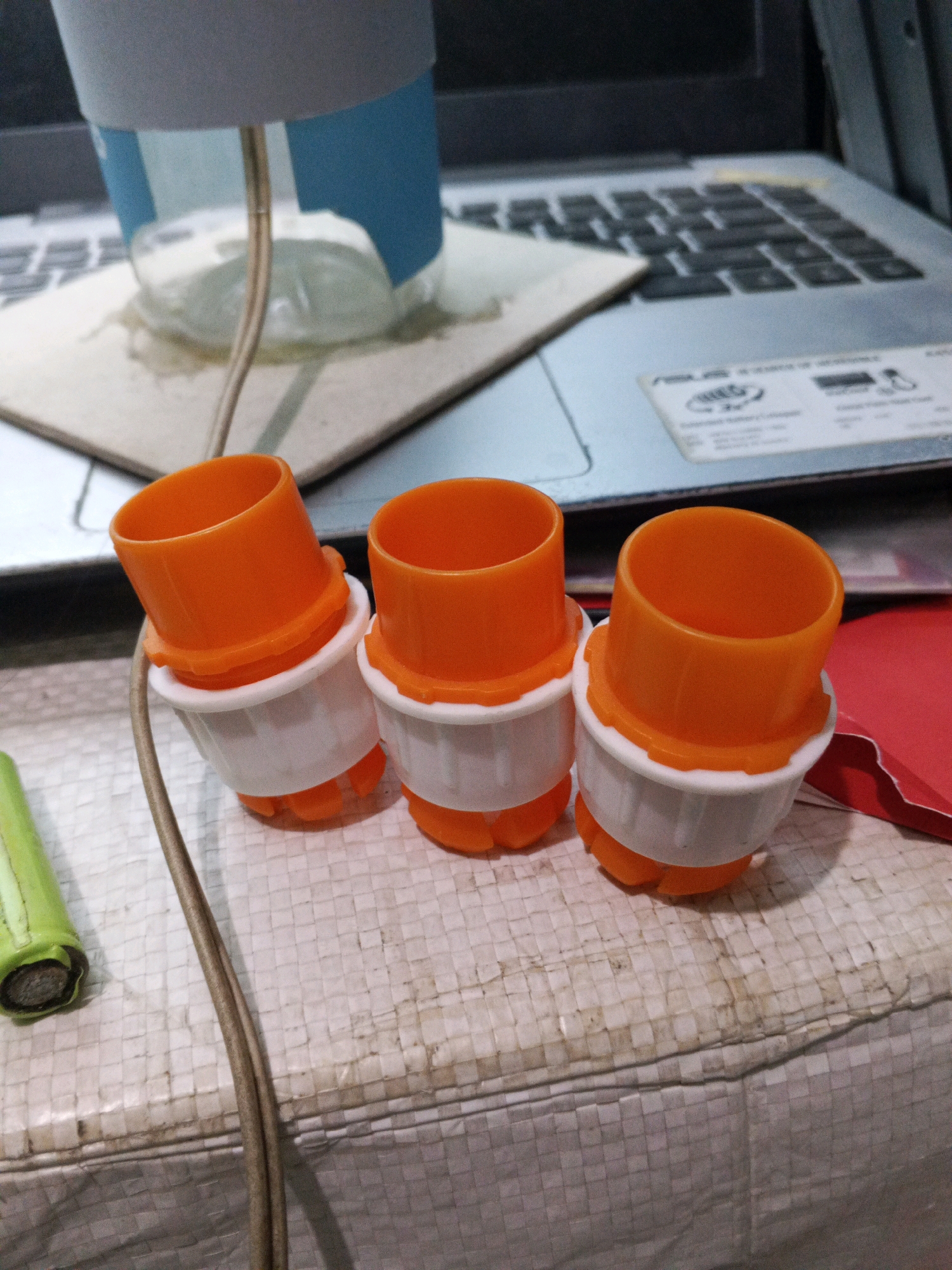 The image shows three orange and white plastic cylindrical objects, possibly kitchen gadgets, arranged in a line on a woven textured surface. In the background, a laptop keyboard and a partially visible lamp with a blue cylinder are present. A green cylindrical object, possibly a battery or a pen, is visible in the lower left corner. The scene appears to be an indoor setting.