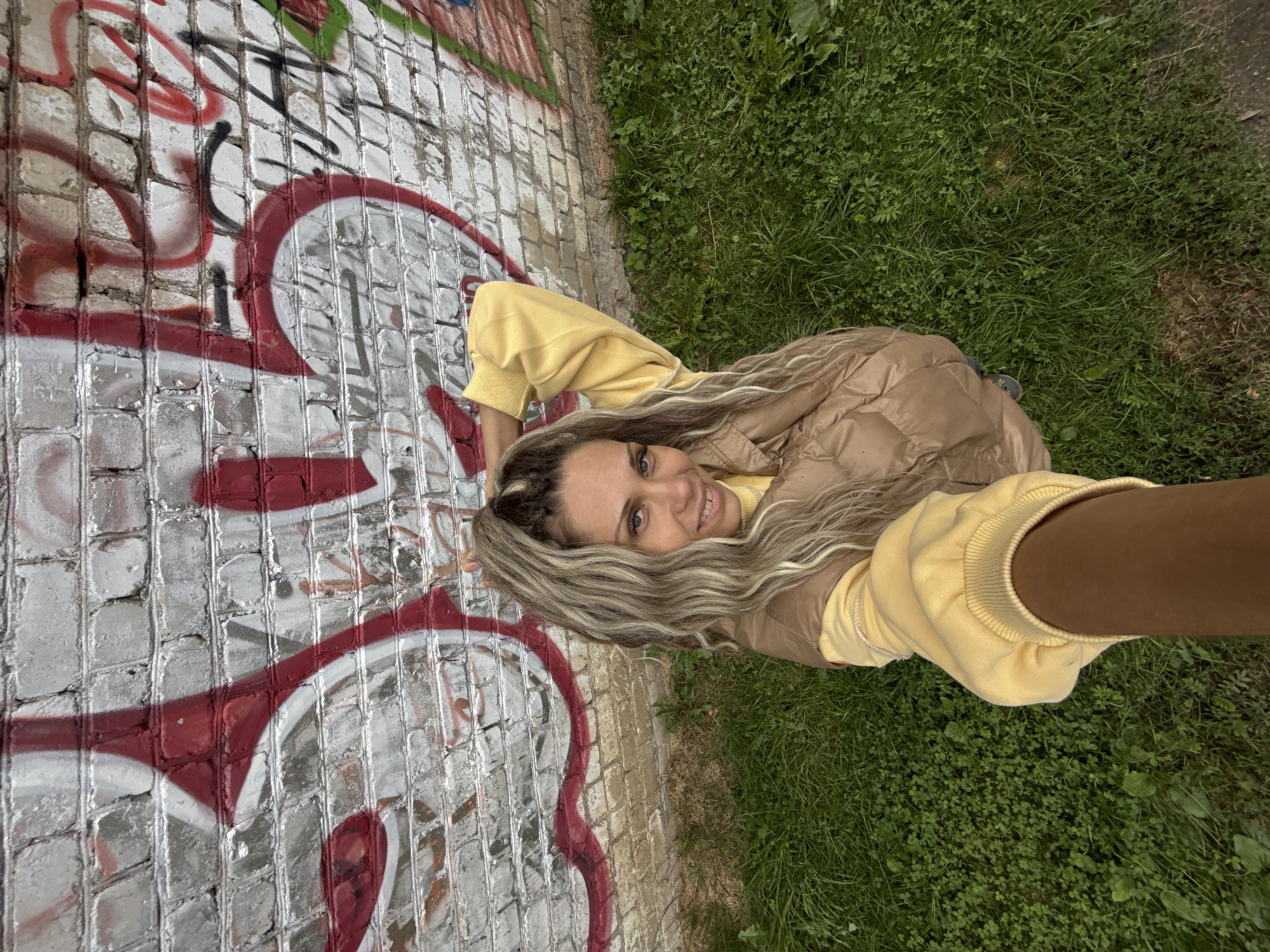 A woman is taking a selfie in front of a graffiti-covered brick wall. She has long, blonde and grey hair that is pulled up into a ponytail. She is wearing a yellow sweatshirt and a brown puffer vest. She is smiling and looking at the camera. The background is a brick wall covered in red and white graffiti. There is also green grass in the foreground.