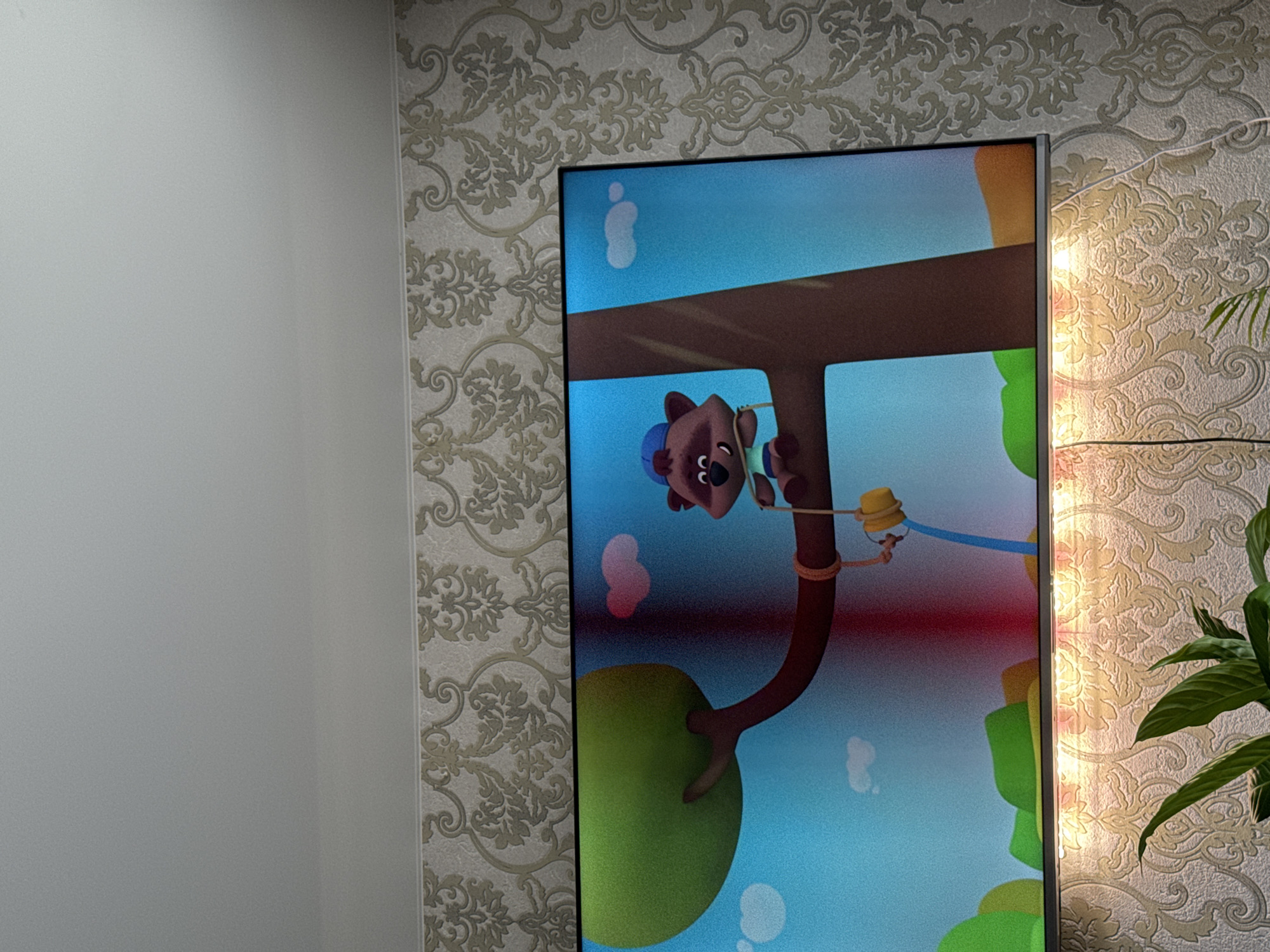 A television displays a cartoon image of a bear hanging from a tree limb, with other cartoon elements in the background. The television is positioned against a wall covered in patterned wallpaper. To the left of the TV is a plain white wall. A plant sits to the right of the TV, partially obscuring the wallpaper. The location is in Chaikovsky, Russia.
