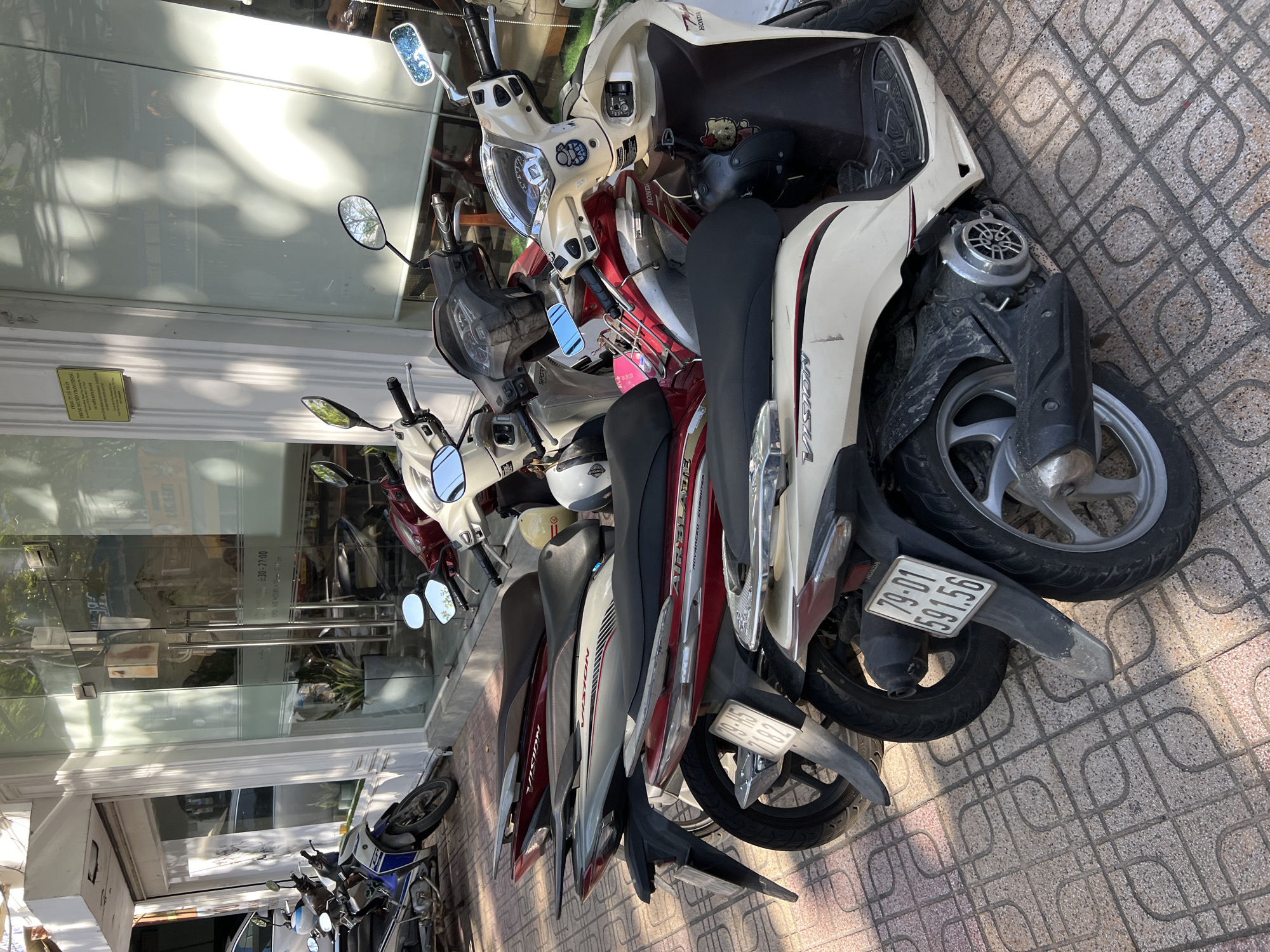 A row of parked scooters is visible on a tiled sidewalk in front of a building in Nha Trang, Vietnam. The scooters are of various colors, including white, red, and blue. Several of the scooters have visible license plates with Vietnamese characters and numbers. The building behind the scooters appears to be a commercial establishment with glass doors and windows, and a sign with text in Vietnamese. Sunlight casts shadows on the sidewalk and the building. One scooter has a "Baby on Board" sticker.