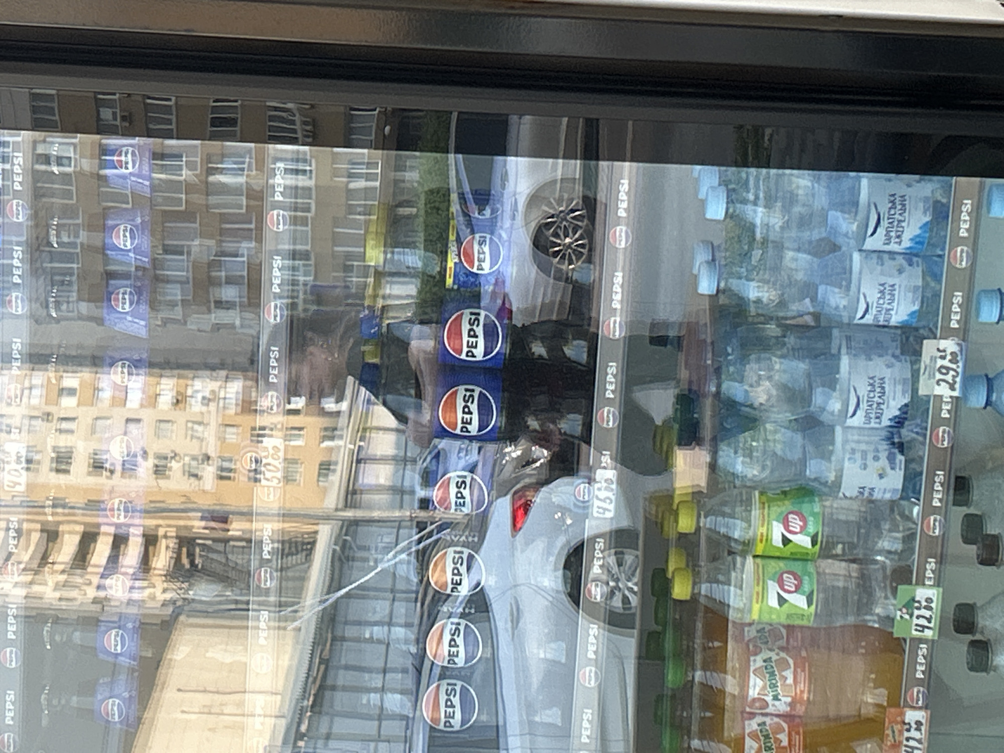 This image captures a reflection of a street scene in a glass-fronted cooler displaying beverages. The cooler is stocked with various bottled drinks, including Pepsi, 7UP, and water bottles labeled "Карпатська Джерельна". Several bottles of Mirinda are also visible. Prominently displayed are multiple Pepsi logos and price tags, with prices like "42.95" and "29.95" visible. In the reflection, a person wearing a dark shirt is partially visible. The background reflection shows a multi-story building with many windows, suggesting an urban environment. The presence of cars in the reflection further confirms this. The overall impression is that of a retail setting, likely an outdoor kiosk or shop, in Brovary, Ukraine, given the Ukrainian labeling on the water bottles.