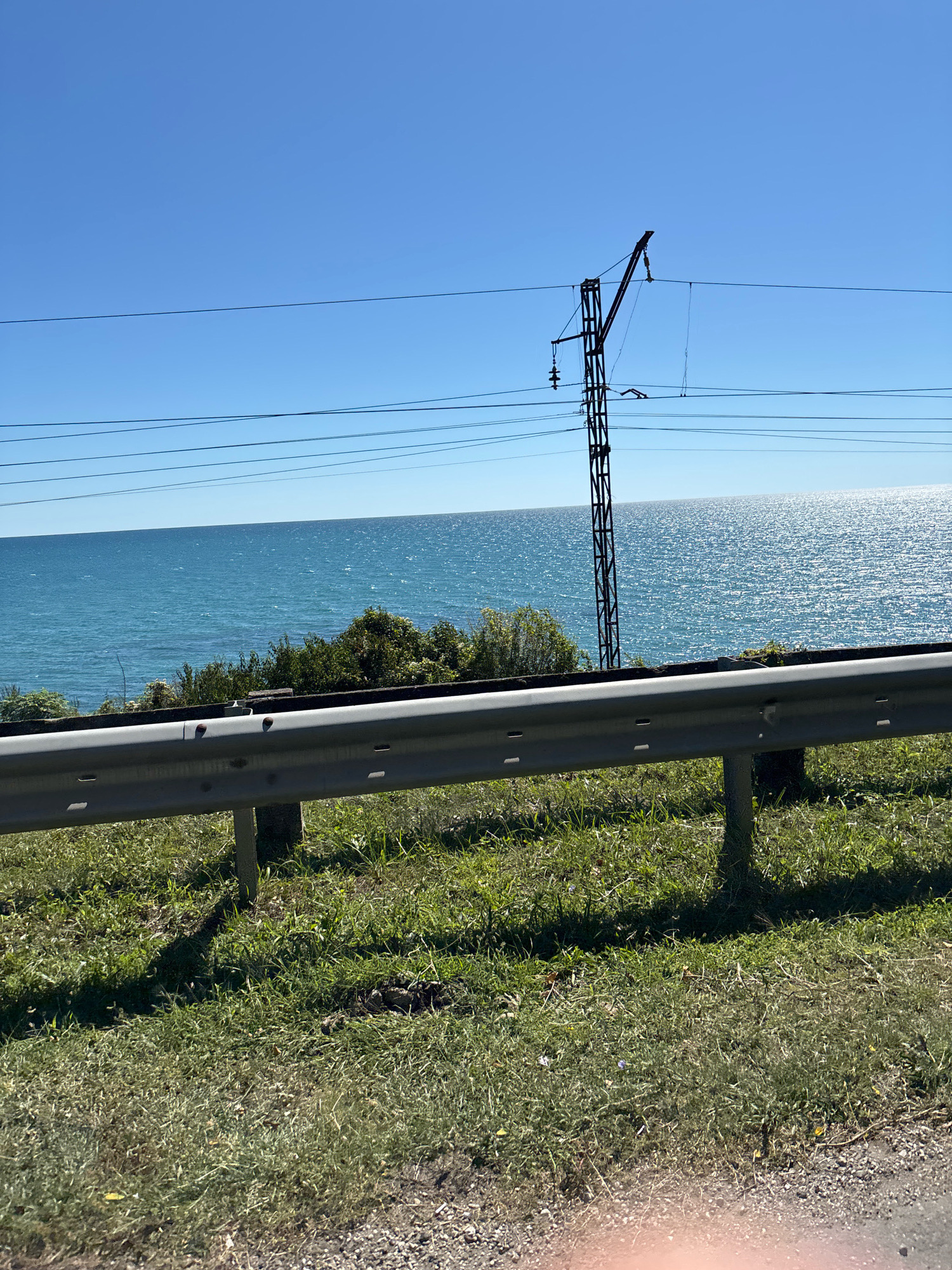 A landscape photograph captures a bright, sunny day with a clear blue sky overhead. In the foreground, a metallic guardrail runs horizontally across the frame, supported by dark posts. Beyond the guardrail, a grassy slope slopes down towards a body of water. The water is a vibrant turquoise, shimmering with sunlight. The horizon is lined with lush green trees and bushes. In the background, a tall, dark utility pole stands against the sky, with several electrical wires stretching out from it. The overall scene is one of natural beauty and a peaceful coastal environment.