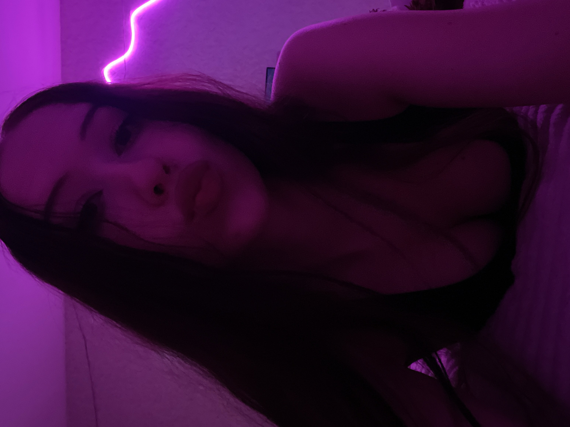The image shows a young woman with long, dark hair, her face partially obscured by shadow and the camera's close proximity. She is wearing a dark top that reveals her cleavage. The lighting is predominantly purple, cast by neon lights that create a wavy line along the wall in the background. The woman appears to be indoors, possibly in a bedroom, with a textured surface, likely bedding, visible in the foreground. The overall mood is intimate and dimly lit. There is no discernible information in the image that would allow for its location to be identified as Votkinsk, Russia.