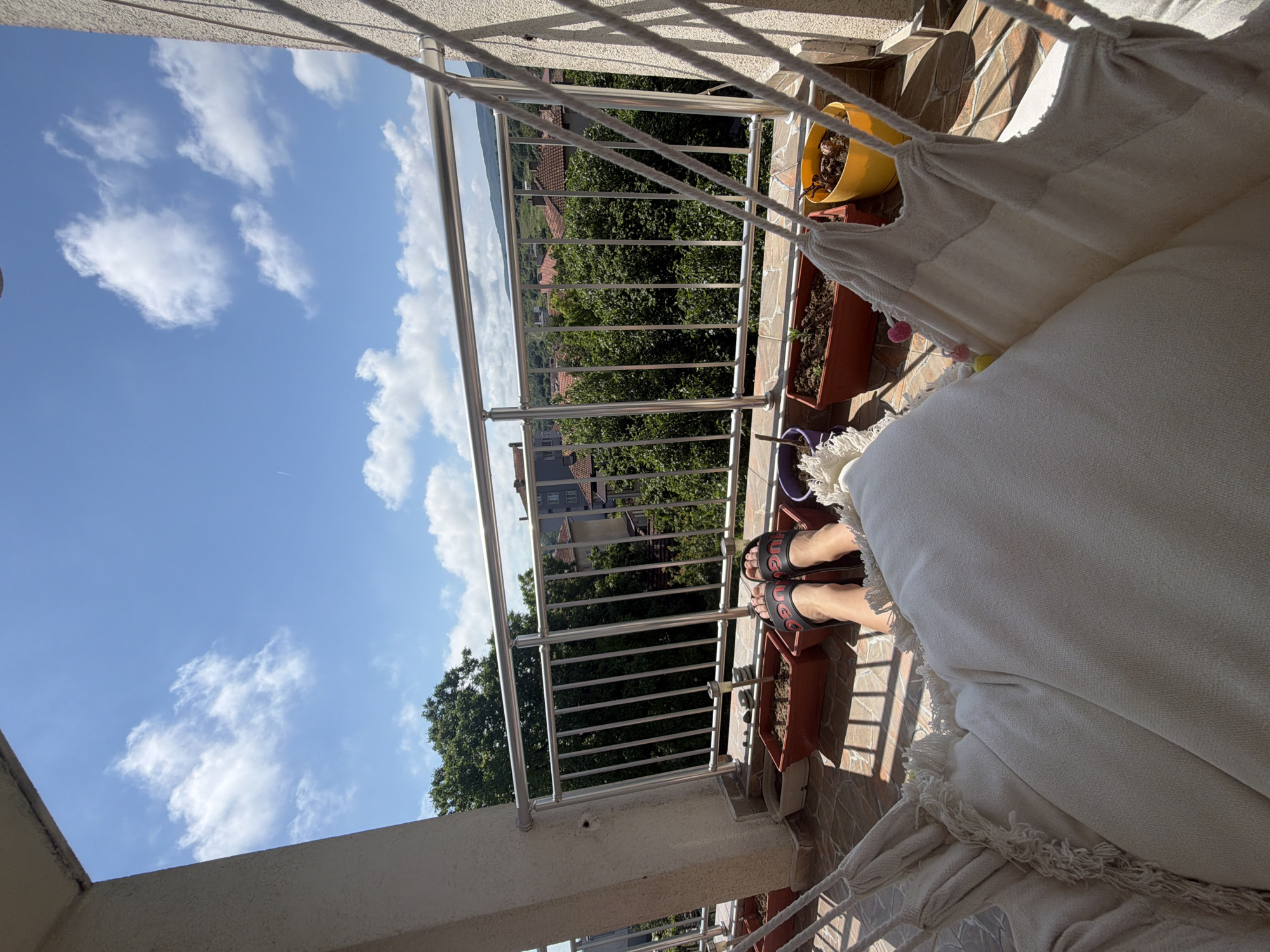 An outdoor perspective shot taken from a balcony in Tsarevo, Bulgaria, shows a person relaxing in a hammock. The hammock, made of off-white fabric with fringed edges, is suspended by thick ropes. The person's legs, clad in black and red slides with a logo, are visible in the foreground, propped up on the hammock's cushion. The balcony floor is tiled with a pattern of beige and terracotta squares. Along the railing of the balcony, several planters are arranged, some containing soil and small plants. A metal railing with vertical bars runs across the balcony, offering a view of a densely forested area dotted with red-tiled roofs of houses. The sky above is a vibrant blue, filled with scattered white cumulus clouds. Sunlight casts distinct shadows on the balcony floor.