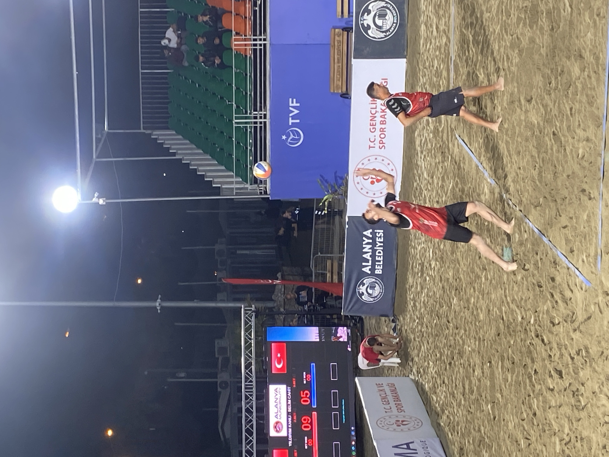 The image shows a beach volleyball game in Alanya, Turkey. Two beach volleyball players are actively engaged on the sand court, one of whom is setting the ball in the air. The score is displayed on a digital scoreboard, indicating 9-5 in favor of "YILDIRIM KANLI - SELIM CAHIT." Various banners and signs surround the court, including logos for "TVF", "ALANYA BELEDİYESİ", and "T.C. GENÇLİK VE SPOR BAKANLIĞI." A row of stadium-style seating can be seen in the background.