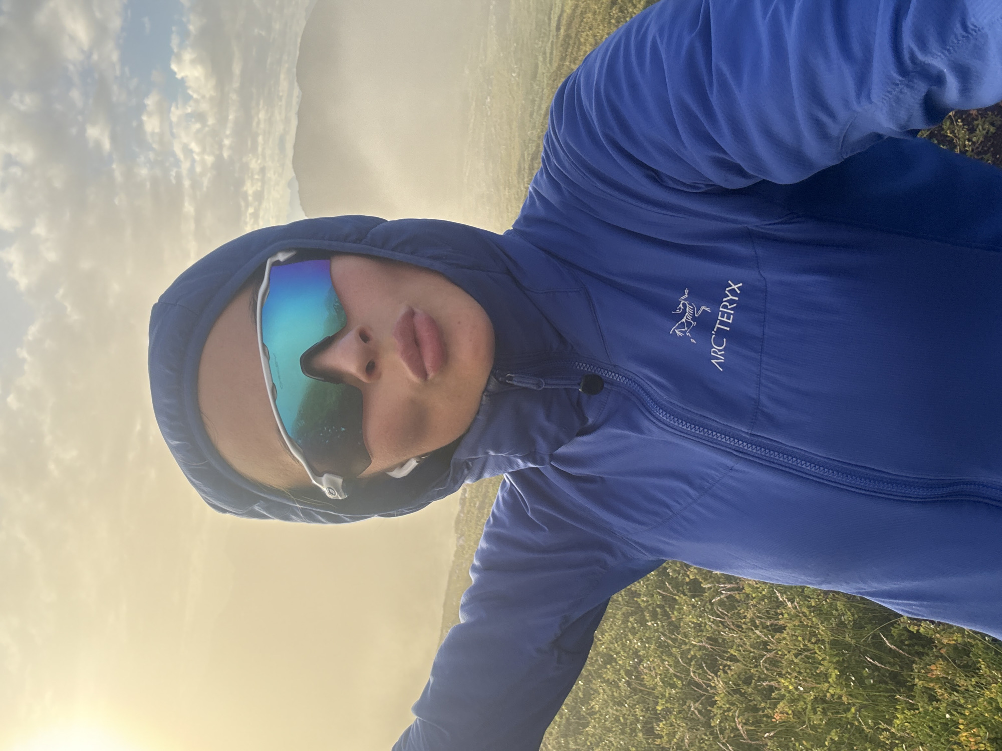 A selfie taken outdoors in a natural environment. A person wearing a blue hooded jacket with the Arc'teryx logo is captured from the chest up. They are also wearing mirrored sunglasses and earbuds, and their lips are pursed. The background features green vegetation, hills or mountains obscured by fog, and a bright sky with clouds, suggesting early morning or late evening light. The image evokes a sense of outdoor activity or adventure. The location appears to be a scenic outdoor area, possibly on the outskirts of Harstad, Norway, given the natural landscape and potential for misty conditions.