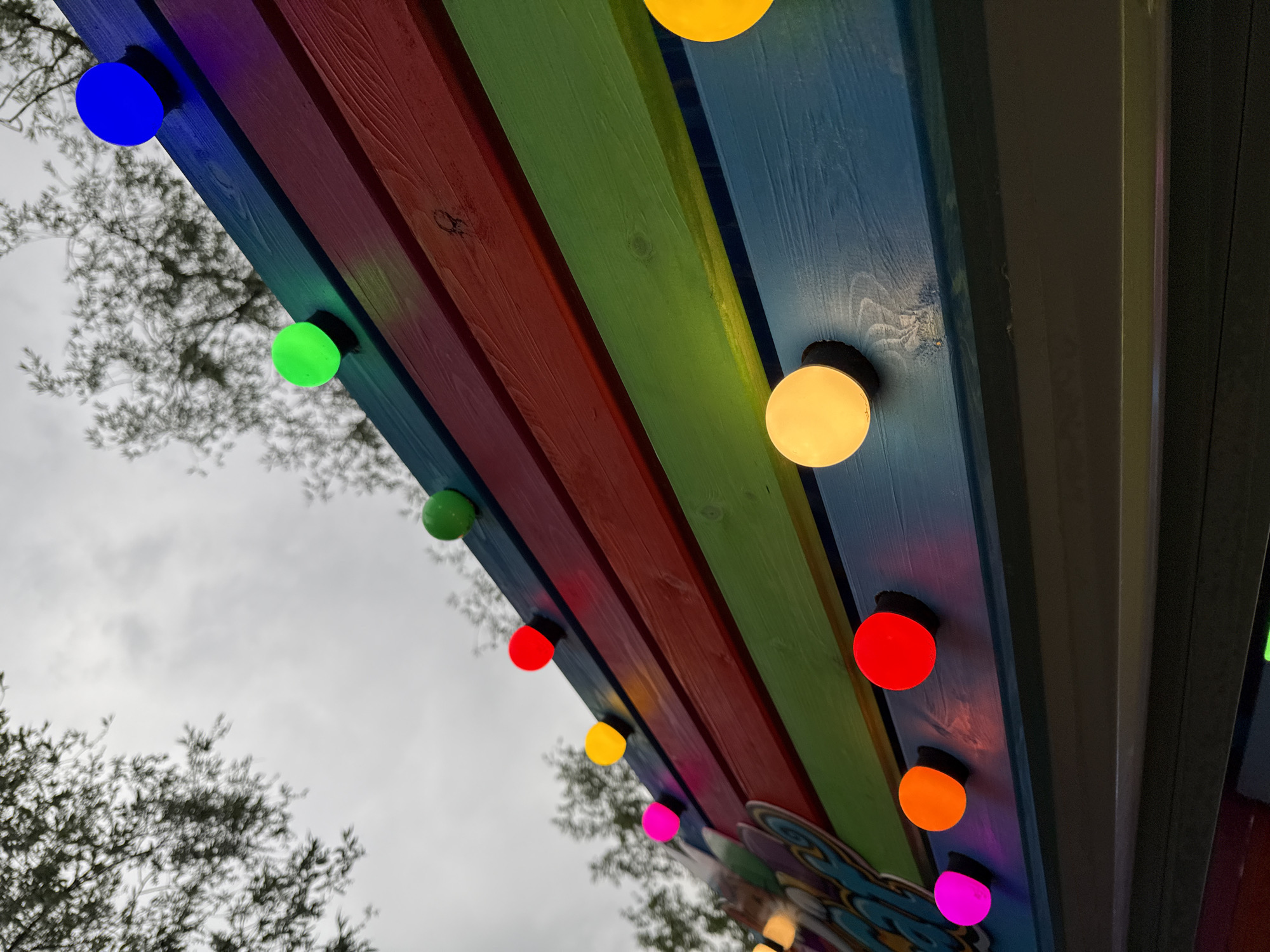 The image shows a low-angle view of a brightly painted awning, possibly part of a building or structure in Chaikovsky, Russia. The awning is constructed of parallel wooden planks painted in a rainbow of colors - blue, green, red, yellow, and pink. Attached to the underside of the awning are numerous round, colored light bulbs. The light bulbs are lit, and their colors include blue, green, red, yellow, orange, and pink. The sky is overcast. Bare tree branches are visible in the lower left and upper left of the frame. The image appears to be taken from a location looking upwards towards the awning.