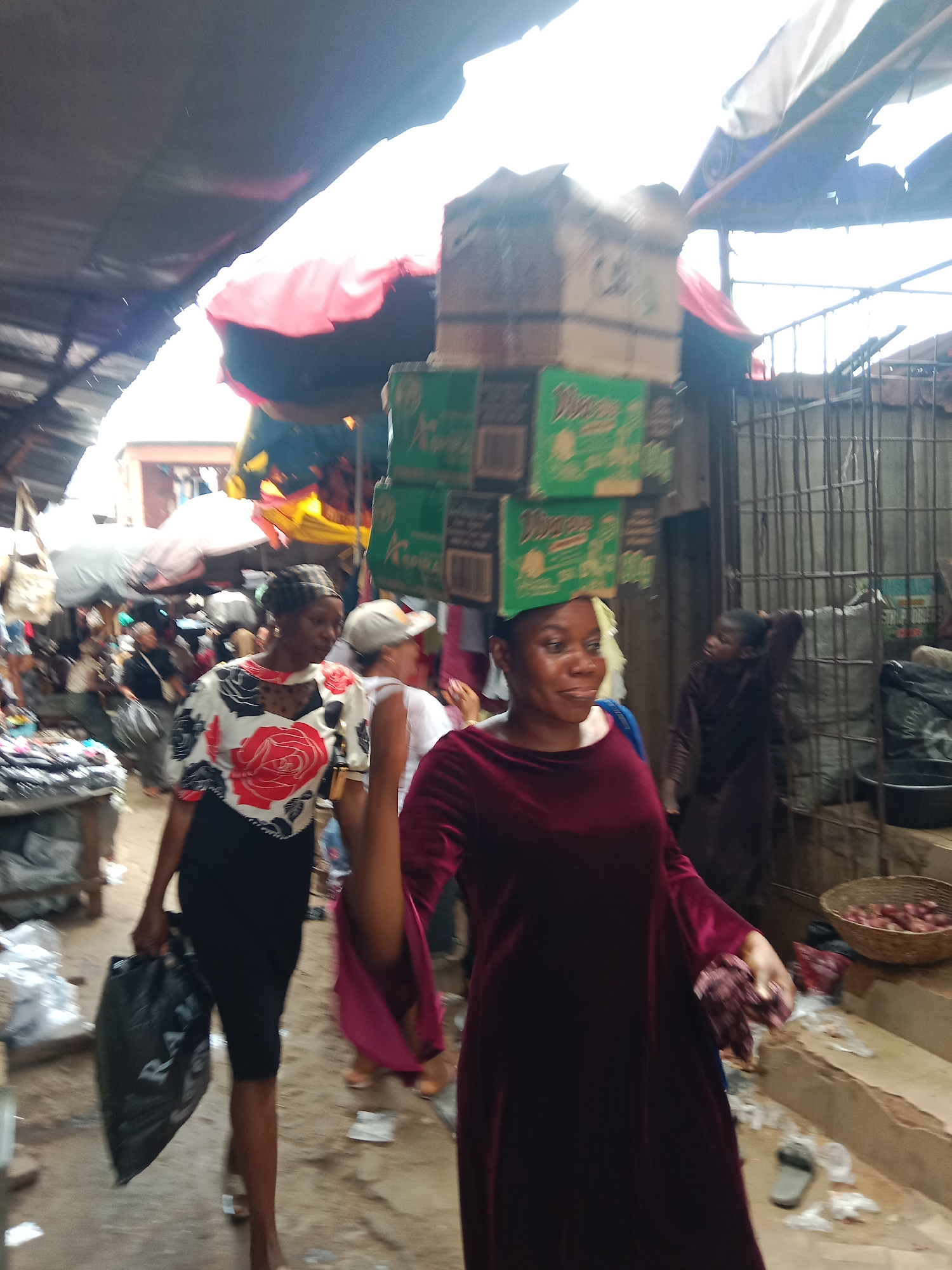 This daytime image captures a vibrant and bustling outdoor market scene in Nkpor, Nigeria. The setting appears to be a narrow thoroughfare lined with makeshift stalls, covered by dark tarpaulin and corrugated metal roofs, through which bright sunlight filters.
In the foreground and central-right, a woman with dark skin is prominently featured, walking and skillfully balancing a stack of approximately seven cardboard boxes on her head. The boxes are a mix of green and brown, with "NIMOS" partially visible on some of the green ones. She is wearing a long-sleeved, dark red velvet dress and has a gentle smile on her face, looking towards the left. Her left hand is slightly raised, holding a piece of dark red fabric matching her dress.
To her left, in the middle ground, another woman walks towards the viewer, carrying a black plastic bag in her right hand, which faintly displays text that might be "WONGA." She wears a short-sleeved top featuring a bold floral pattern of red roses and black leaves on a white background, paired with a black skirt or shorts, and a dark, patterned headscarf.
The background is filled with blurred figures of other people, suggesting a crowded and active environment. To the far right, a younger person or child in a dark outfit is visible behind dark metal bars or a gate. Further details of the market include various goods, such as a woven basket filled with red onions or shallots placed on a raised platform on the right. The ground is dirt, with scattered debris and a pair of slippers. The scene conveys the daily life and commerce typical of a lively market.