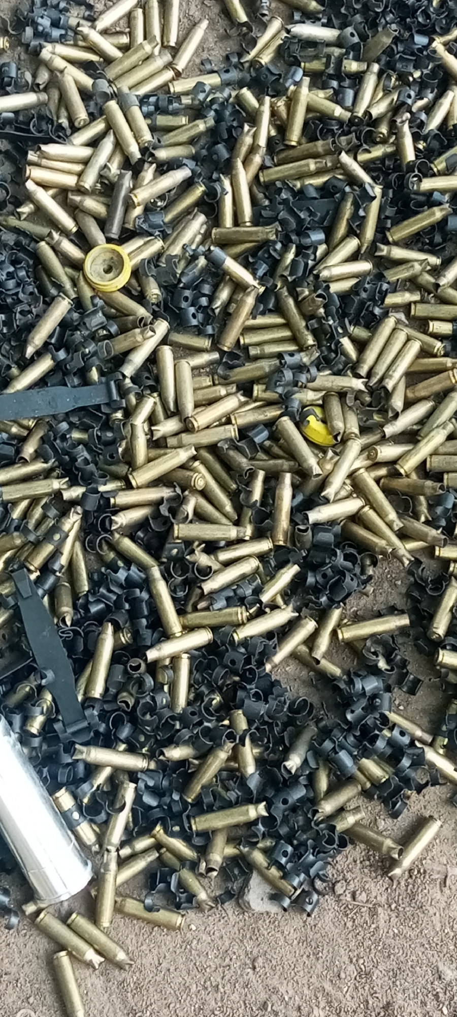 The image shows a collection of spent brass bullet casings and black metal cartridge links scattered on a dirty, possibly concrete surface. The casings and links are mixed together. The location of Adankolo, Nigeria is extraneous to the image and is not discernible from the visual information.