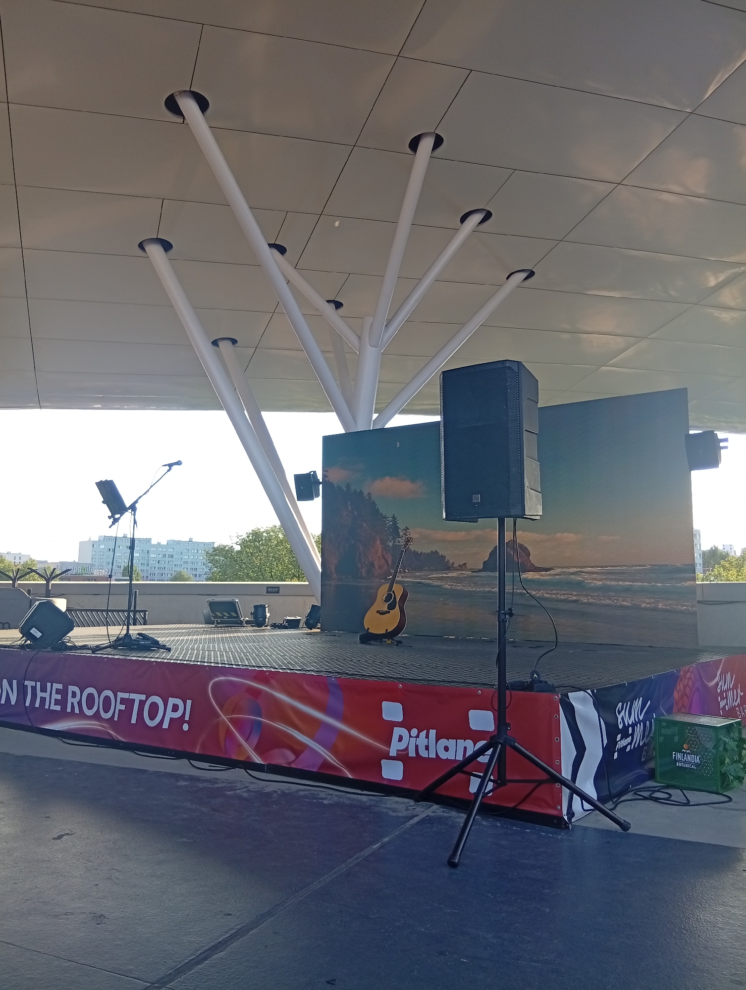 This image depicts an outdoor stage area set up for a performance in Wrocław, Poland, likely on a rooftop given the banner text "ON THE ROOFTOP!". The stage itself is elevated and has a black, perforated surface. A large speaker is mounted on a stand to the right of the stage, facing forward. Behind the stage, there is a large digital screen displaying a scenic image of a rocky coastline with waves. A light-colored acoustic guitar rests on the stage near the screen. A microphone stand with a microphone is positioned to the left of the stage. Several other speakers and audio equipment are visible on the stage. The stage is framed by a banner that wraps around its front and sides, featuring colorful graphics and text, including "Pitlan" and "FINLANDIA BOTANICAL". The overhead structure, supported by white, tree-like pillars, suggests a modern architectural design. In the background, apartment buildings and greenery are visible, indicating an urban setting. The overall scene suggests a prepared setup for a live music event.