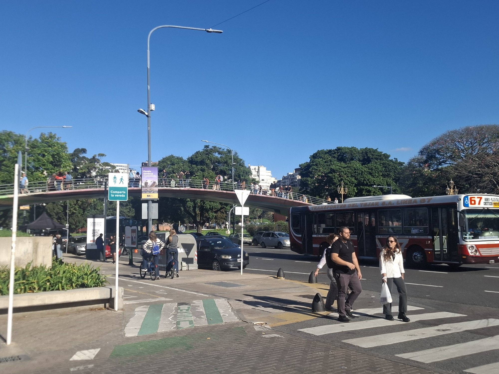 The image depicts a street scene in Buenos Aires, Argentina, on a clear day. Pedestrians are crossing the street at a zebra crossing, and two cyclists are on the sidewalk. A red and white city bus, line 67, is present in the background. Overhead, a pedestrian bridge spans the road, with people visible walking on it. The environment includes trees, streetlights, and several parked cars and buildings in the distance. Street signs are visible with Spanish writing. The image is sharply focused.