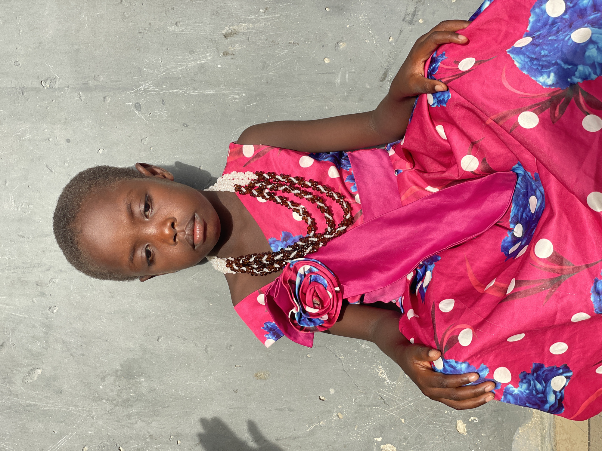 A young, dark-skinned child lies on a textured gray surface, possibly concrete. The child is wearing a bright pink dress adorned with white polka dots and blue floral patterns. A pink satin sash is tied around the child's waist, with a decorative flower detail. The child is wearing a multilayered necklace, with a white pearl layer close to the neck and a brown beaded layer. The child's head is positioned at the top left of the frame. The image shows the child from an angled overhead view, with the child's body at a diagonal across the frame. This could be a place in Badagry, Nigeria.